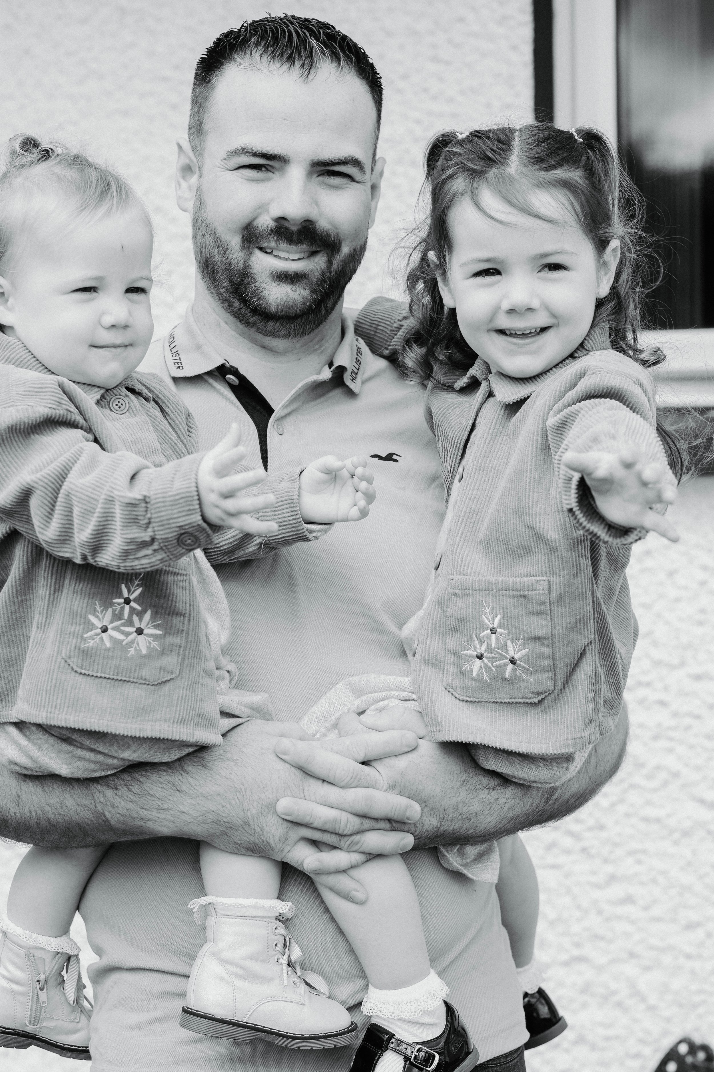 A man holding two young girls, all smiling and looking at the camera. The girls are wearing jackets, and the man has a beard and short hair. The photo is in black and white.