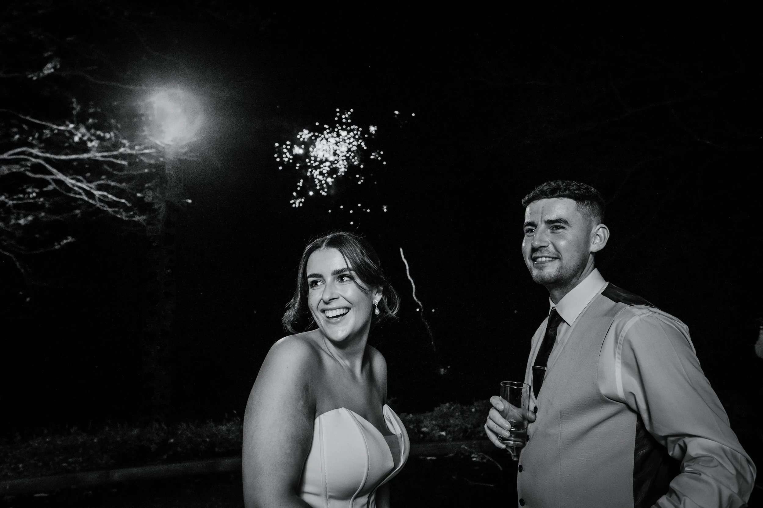 A black-and-white photo of a smiling woman and man celebrating at night with fireworks in the sky, the woman wearing a strapless dress and the man holding a drink.