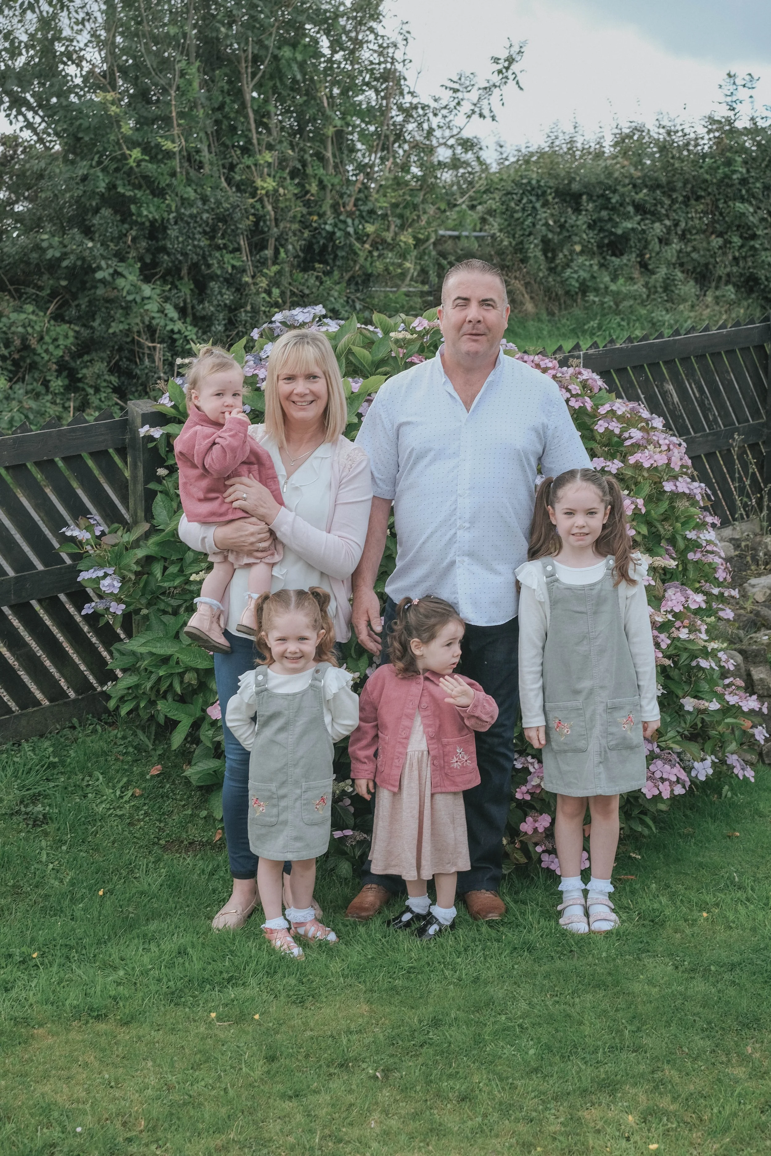 A family of six posing outdoors in front of flowering bushes, with three women and three children, all smiling and standing on grass.