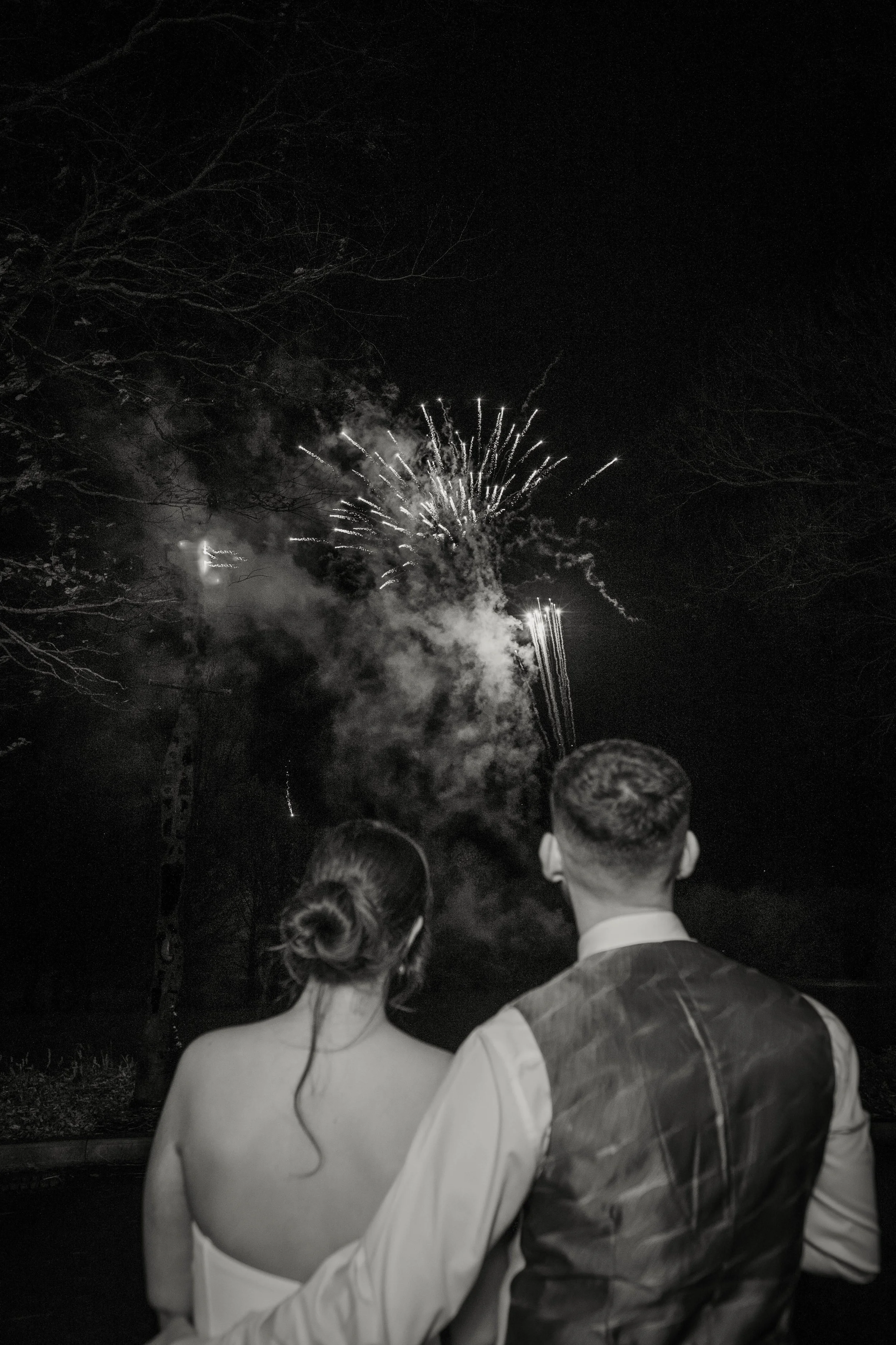 A couple embraces while watching wedding fireworks light up the night sky, seen from behind.