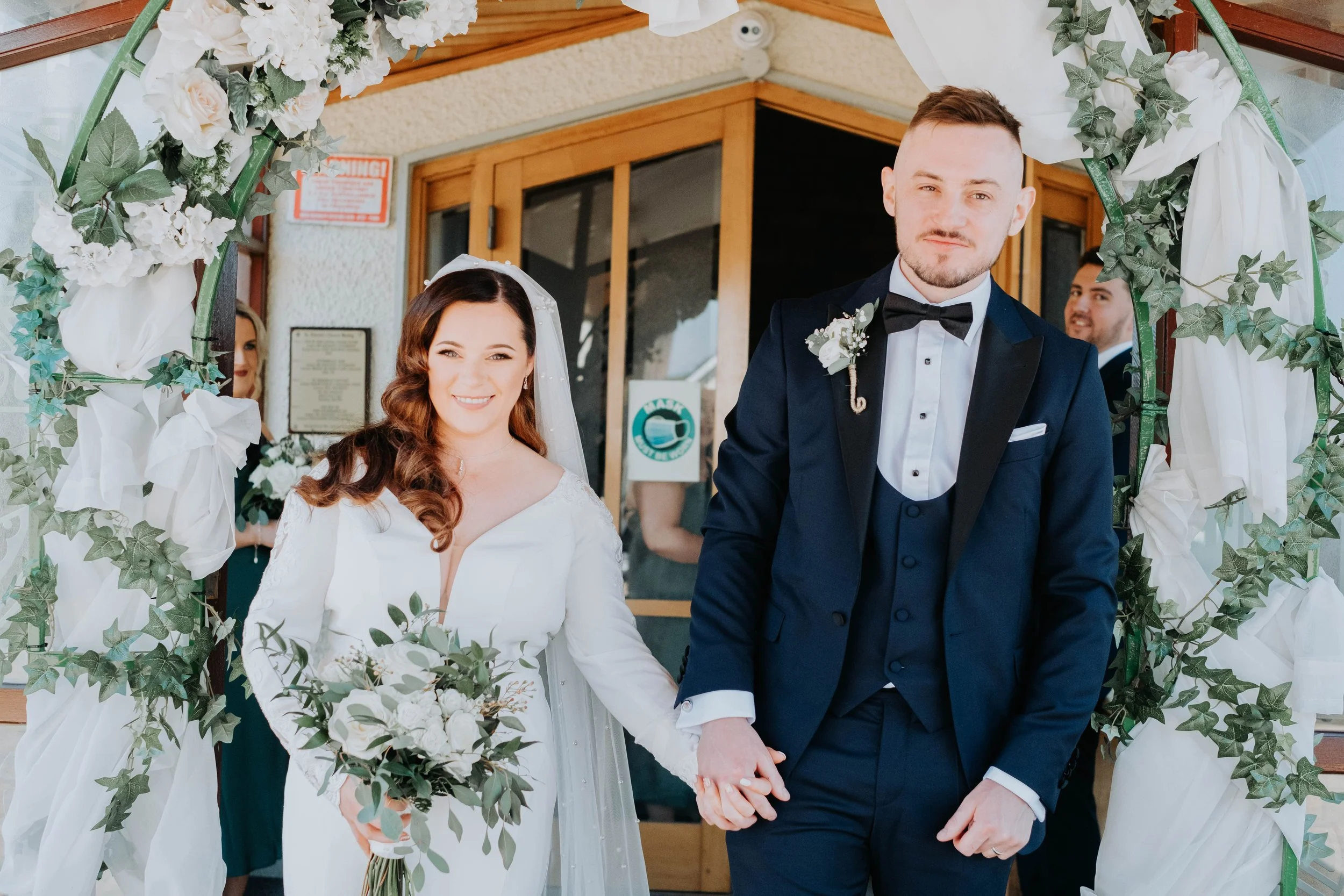 A bride and groom holding hands under a decorated arch at their wedding ceremony. The bride has long, wavy brown hair and the groom has short light brown hair.