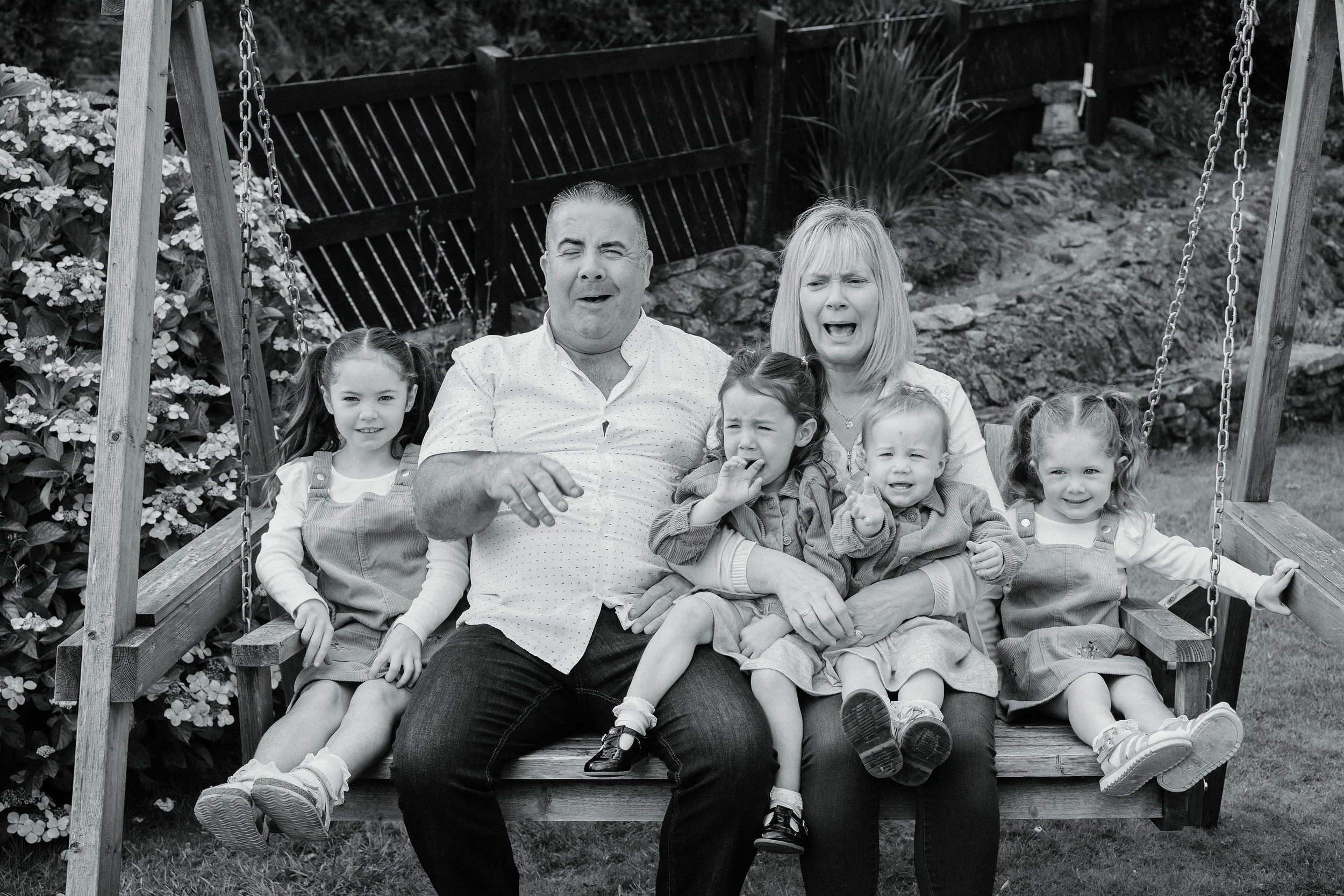A family of 6 with two adults and 4 children on a wooden swing in a garden, all with surprised or distressed expressions.