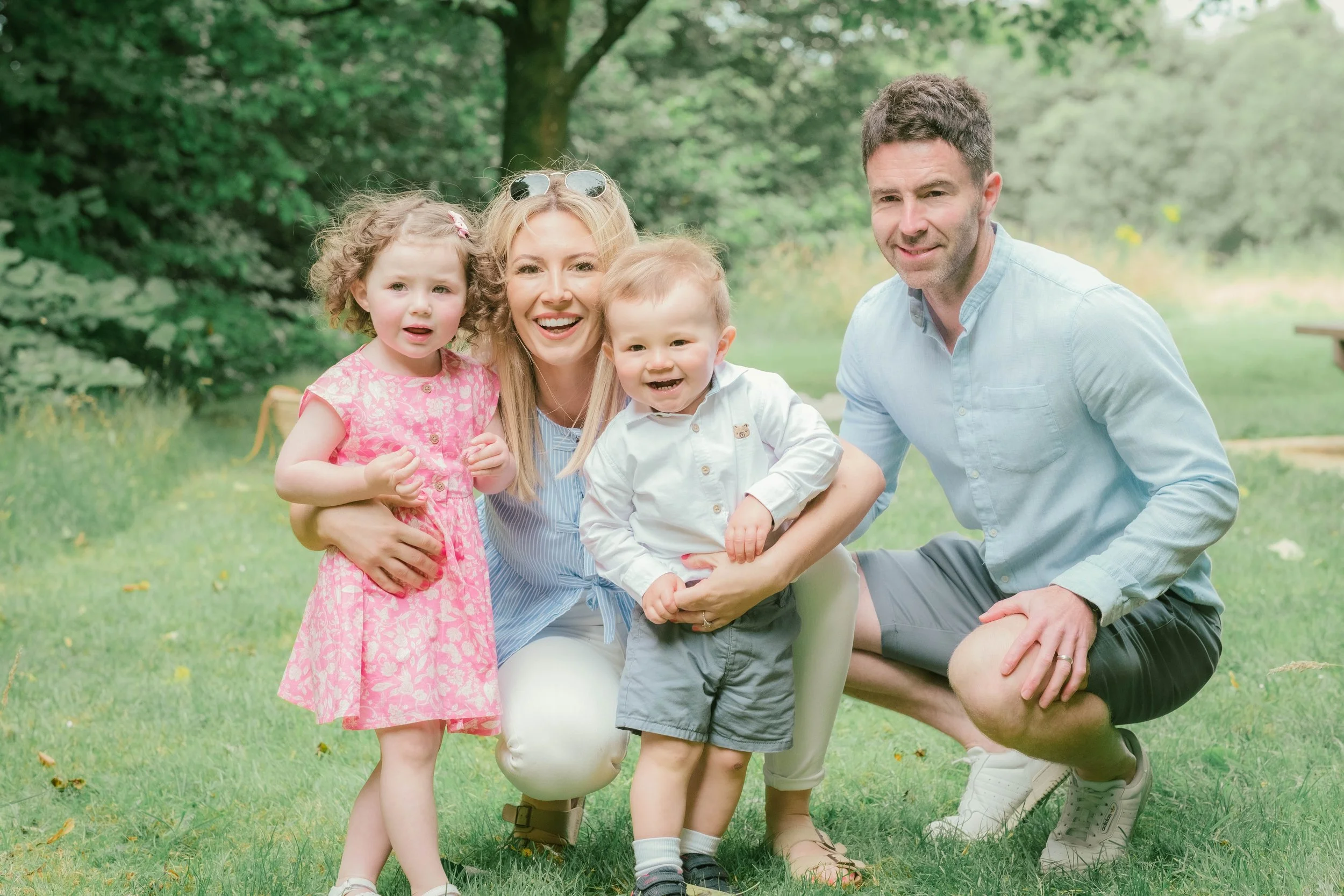 Family of four outdoors on grass, smiling and posing for the camera, with trees in the background. The mother has blonde hair and is holding a young girl in a pink dress. The father has dark hair, wearing a light blue shirt, and is kneeling next to a