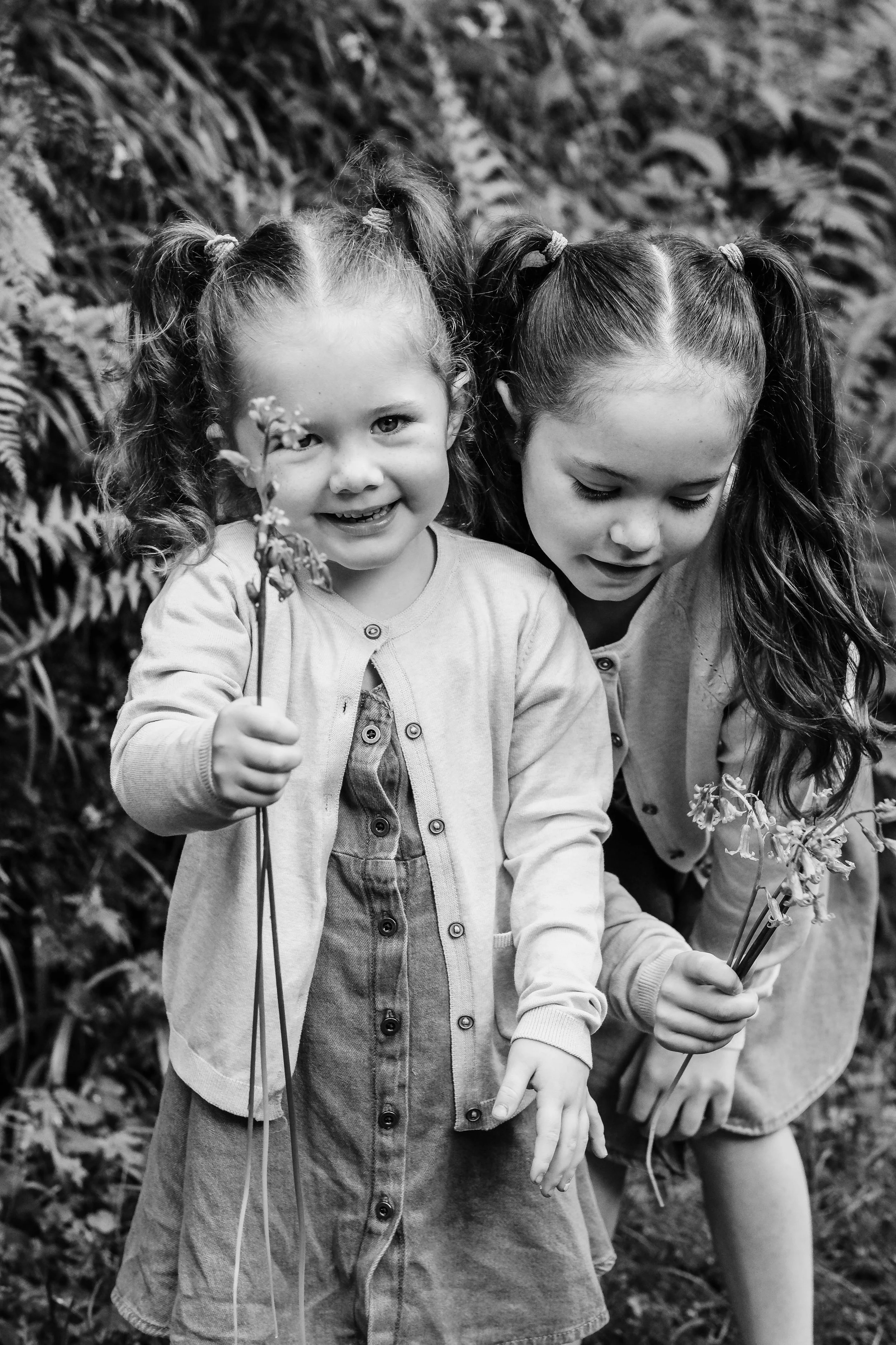 Two young girls with pigtails smiling and holding flowers outdoors in a natural setting, in black and white.