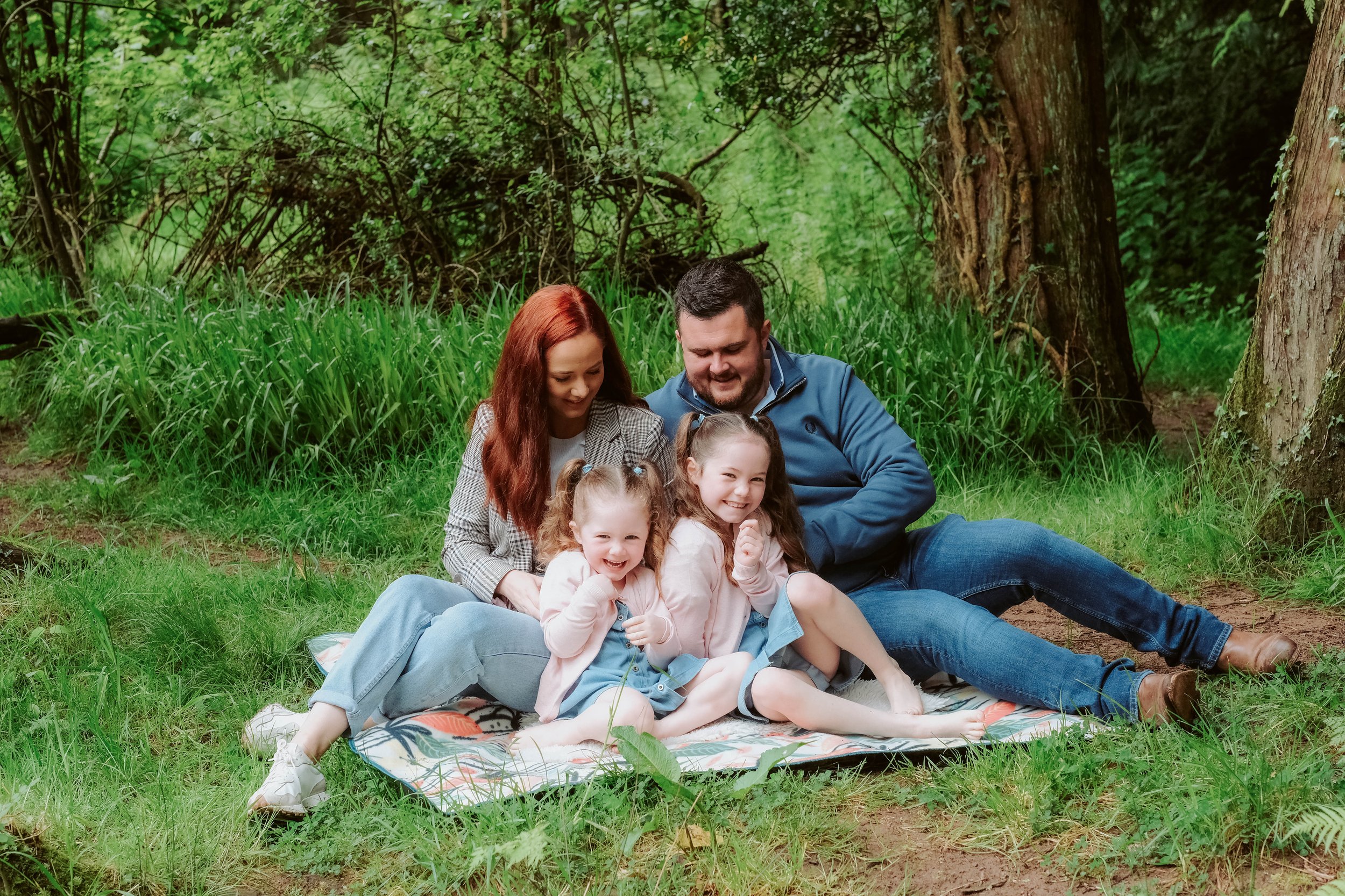 A family of four sitting on a picnic blanket in a green forest, laughing and enjoying the outdoors.