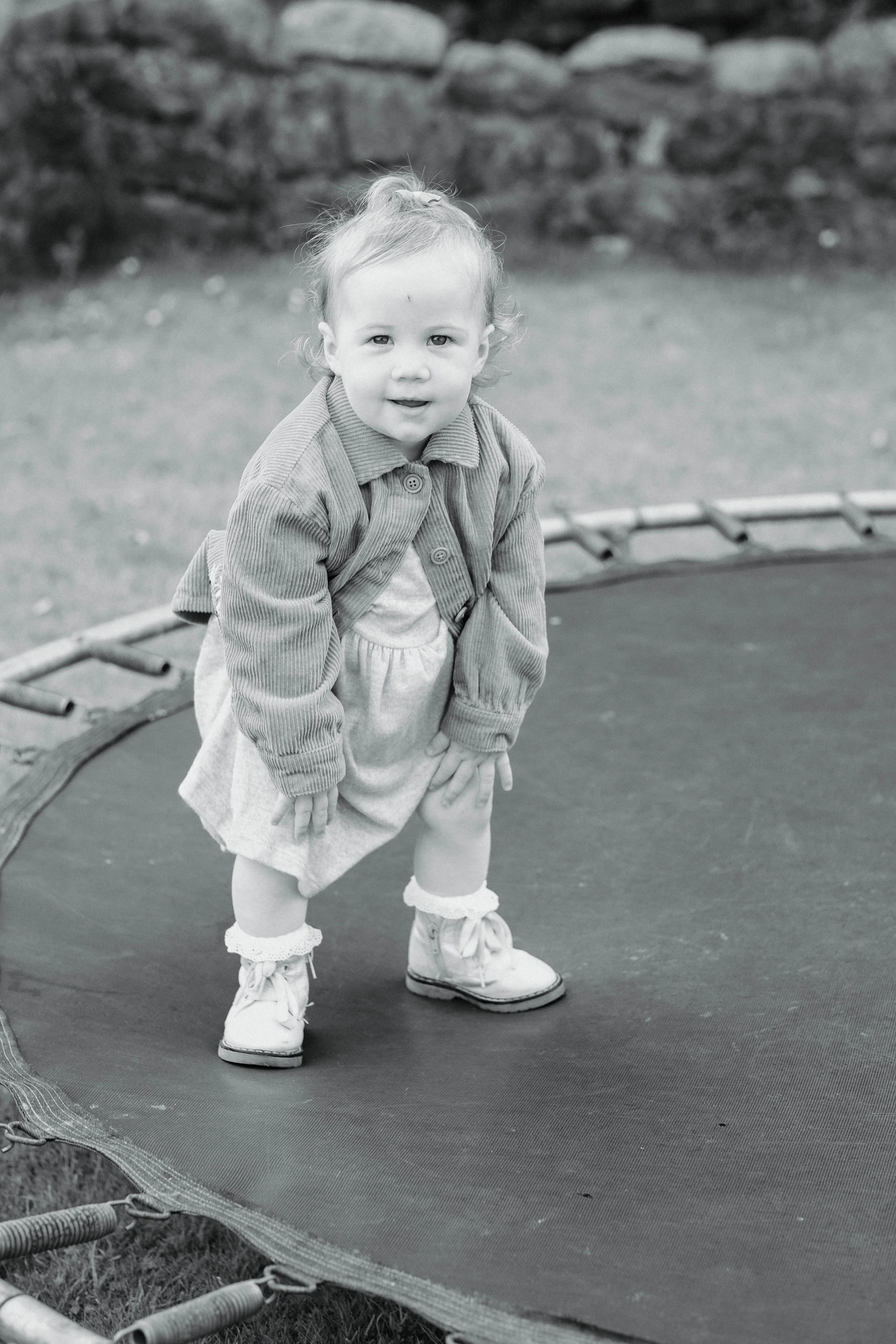 A young girl standing on a trampoline outdoors, dressed in a dress, jacket, socks, and shoes, looking at the camera.