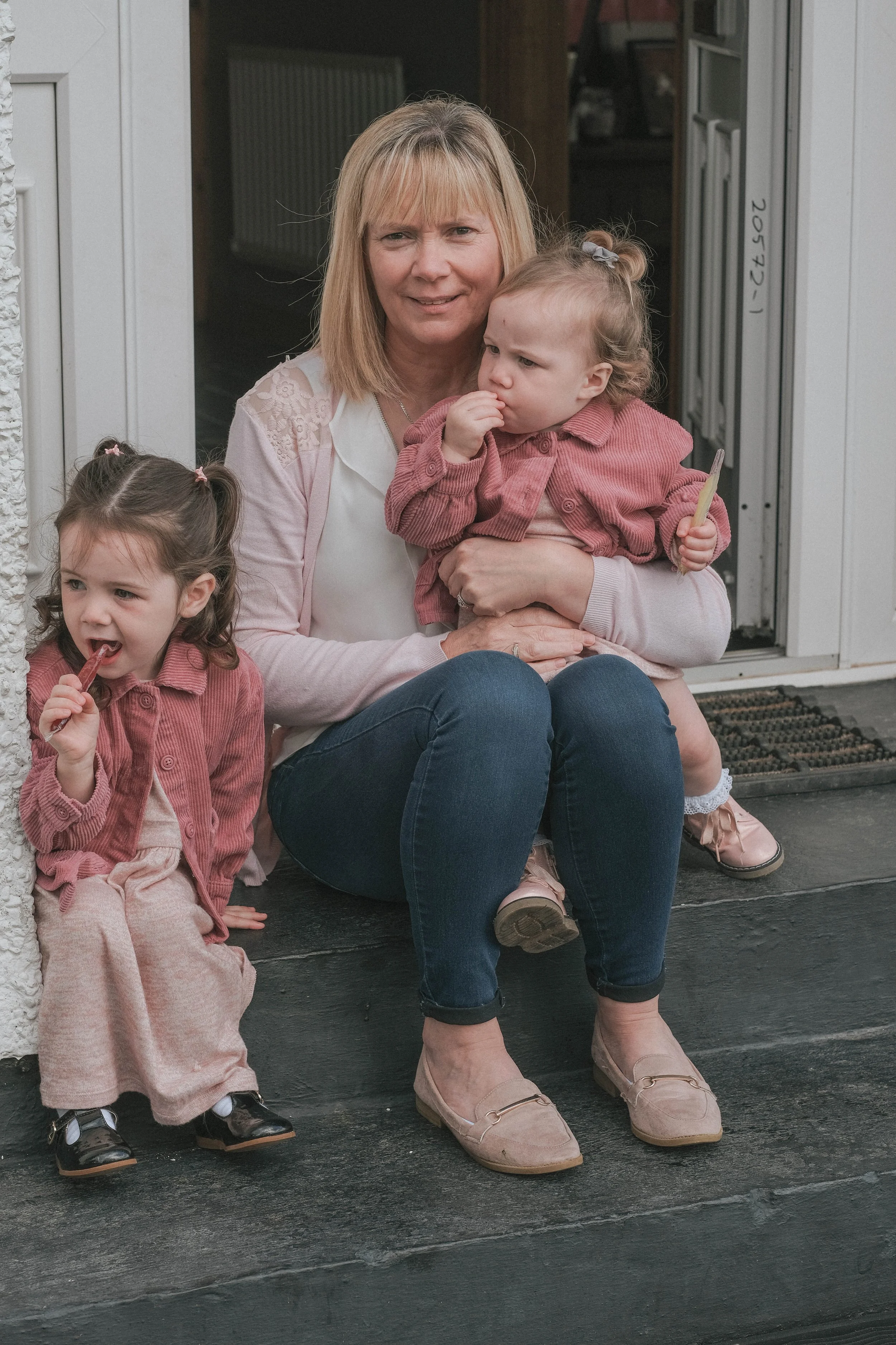 A woman sitting on the front porch with two young girls, one on her lap and one sitting beside her, all wearing pink jackets, with the woman holding a popsicle and the girl on the porch eating a popsicle.