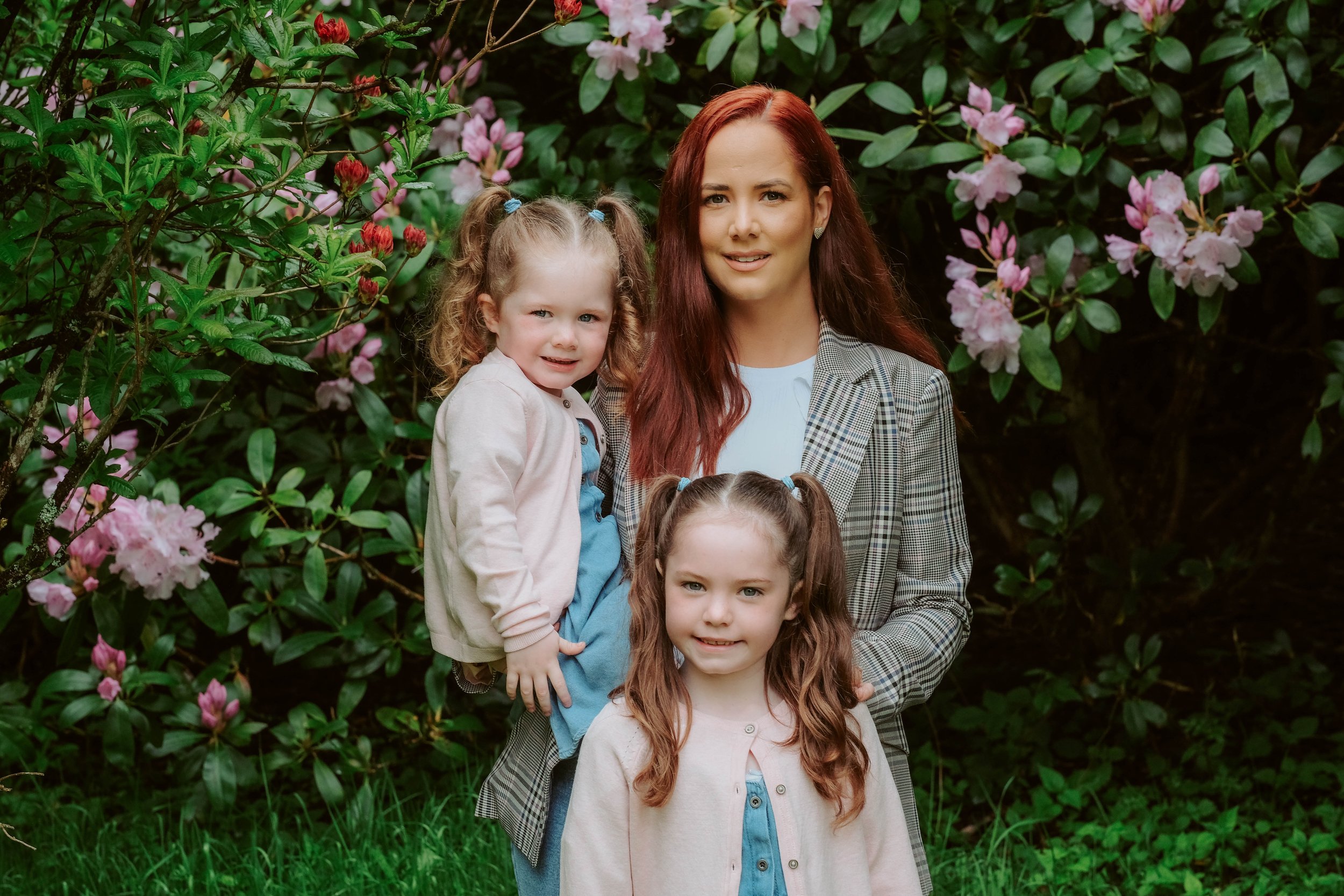 A woman with red hair stands outdoors with two young girls, surrounded by blooming pink flowers and green foliage.