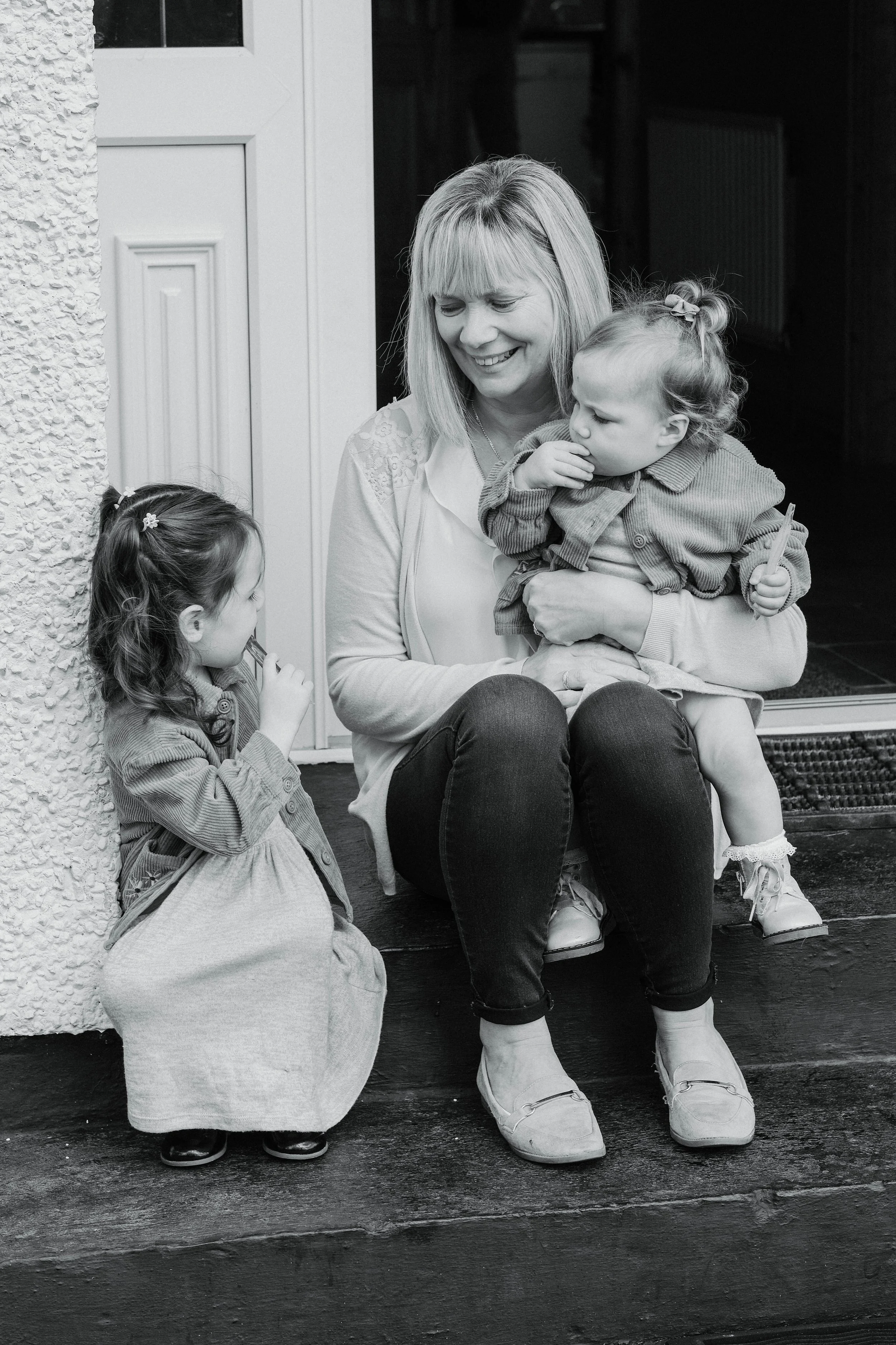 A woman sitting on a wooden porch steps smiling and holding a young girl in her lap, while another girl is sitting on the ground next to her, near an open door.