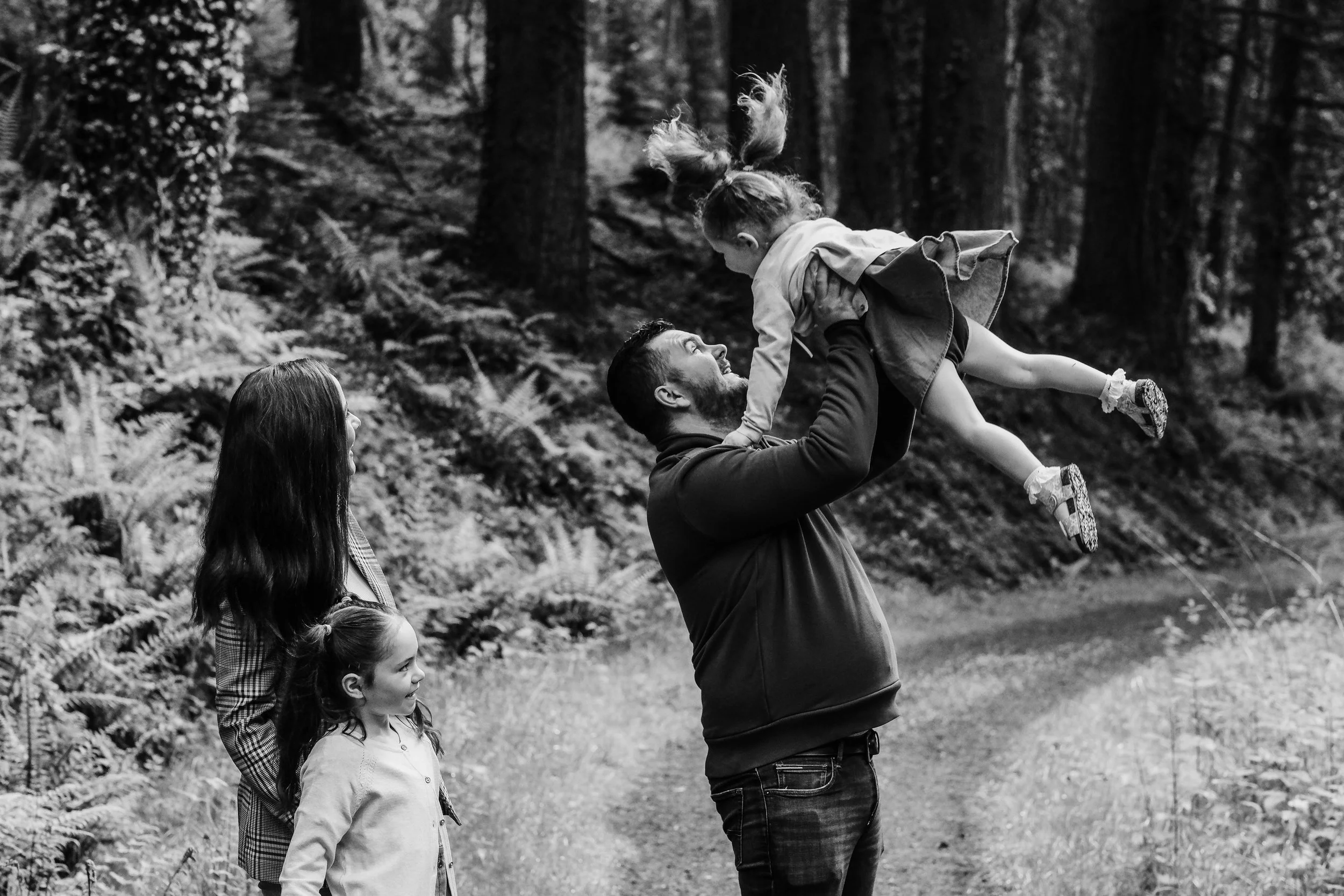 A man lifts a young girl into the air in a forest, with two women and another girl watching, all smiling.