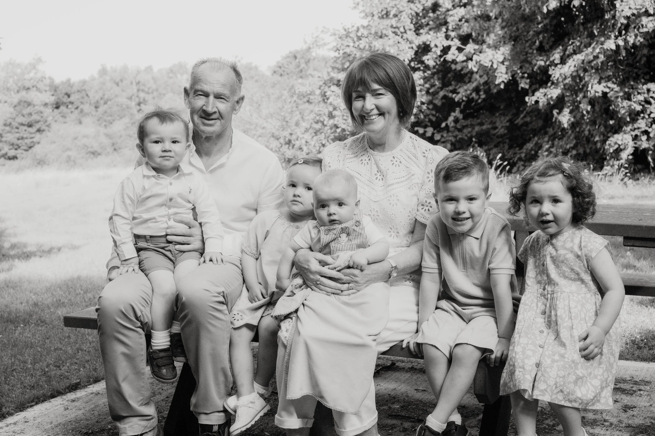 A black and white photo of a family of seven, including two elderly adults and five young children, sitting on a park bench in an outdoor setting with trees in the background.