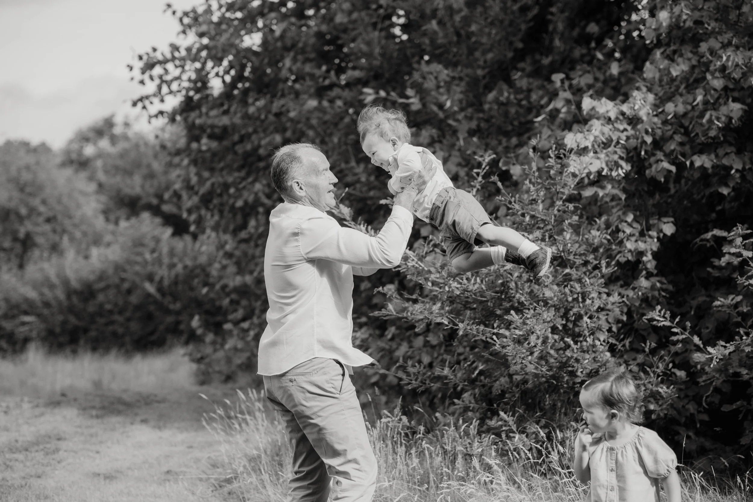 An man playfully lifting a young boy into the air outdoors, with a young girl watching, surrounded by trees and grass.