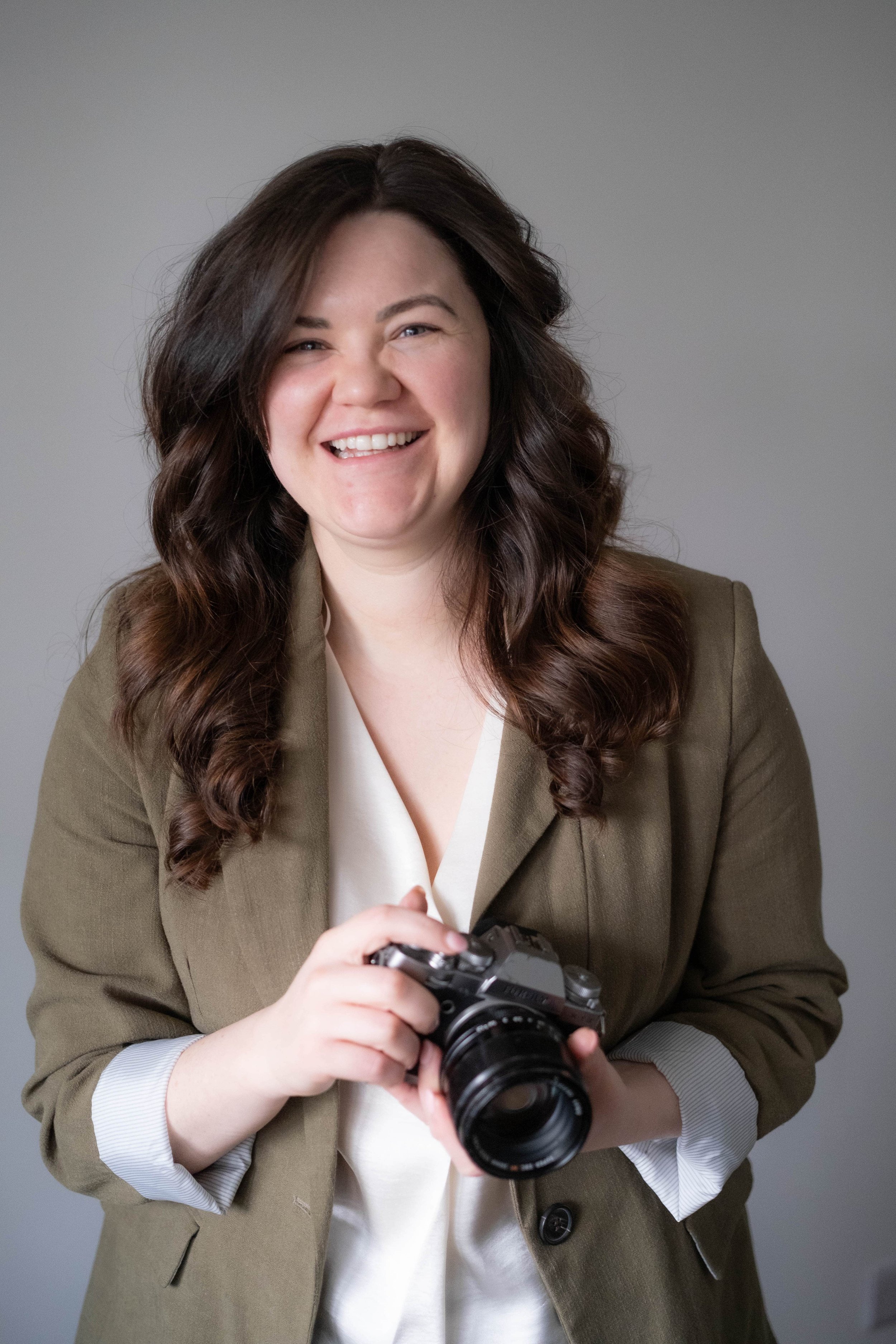 A smiling wedding photographer with long brown hair holds a camera, wearing a brown blazer over a white top against a plain background.