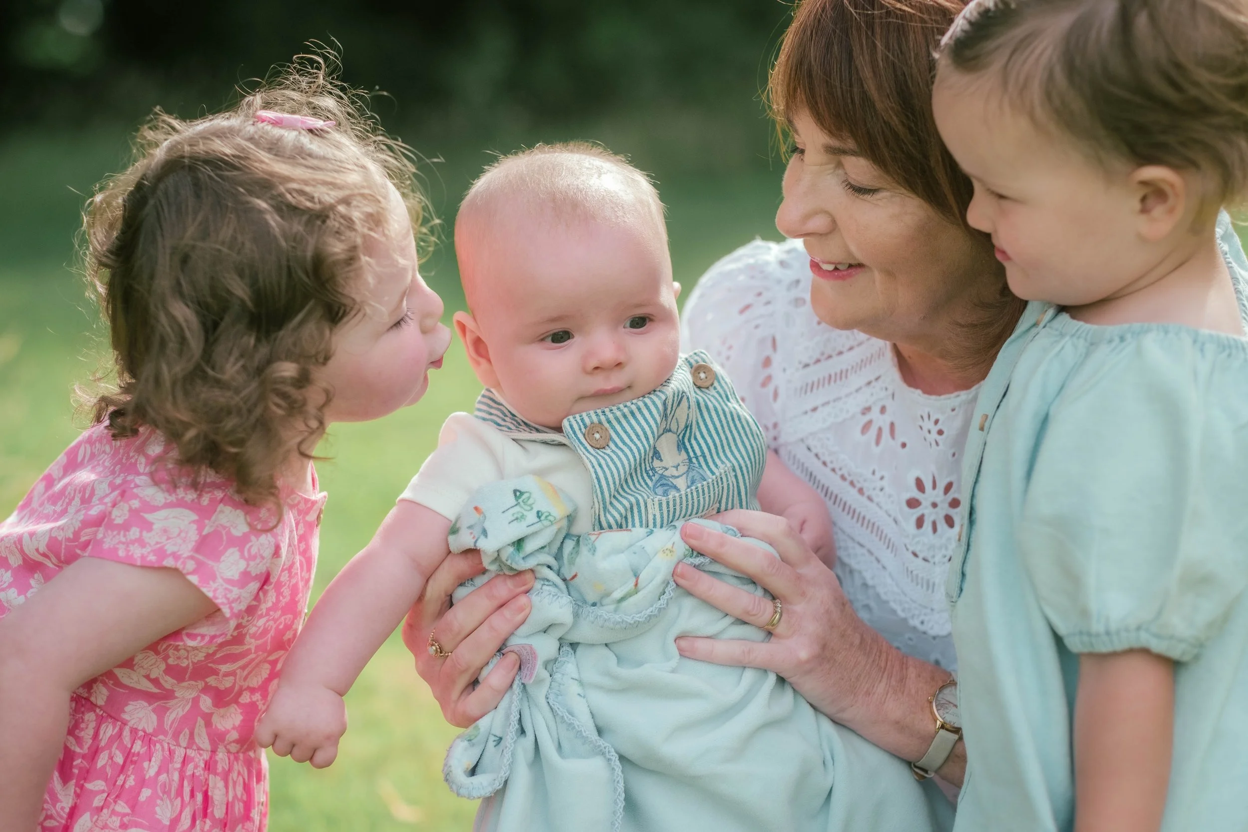 A woman holding a baby boy while two young girls look at him affectionately outdoors.
