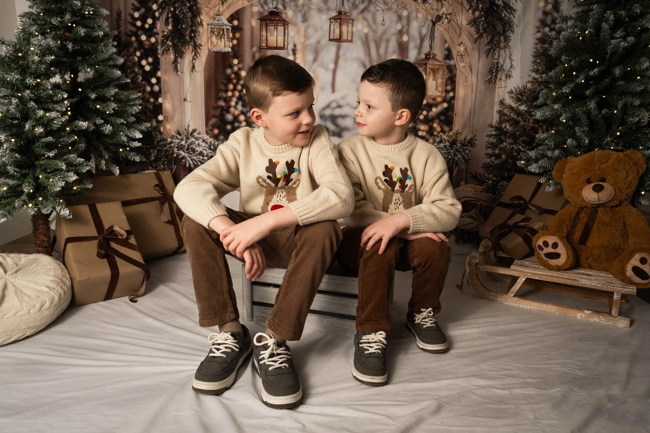 Two young boys sitting closely together on a small bench surrounded by Christmas decorations, including pine trees, wrapped gifts, and a teddy bear on a sled, in a festive holiday setting.