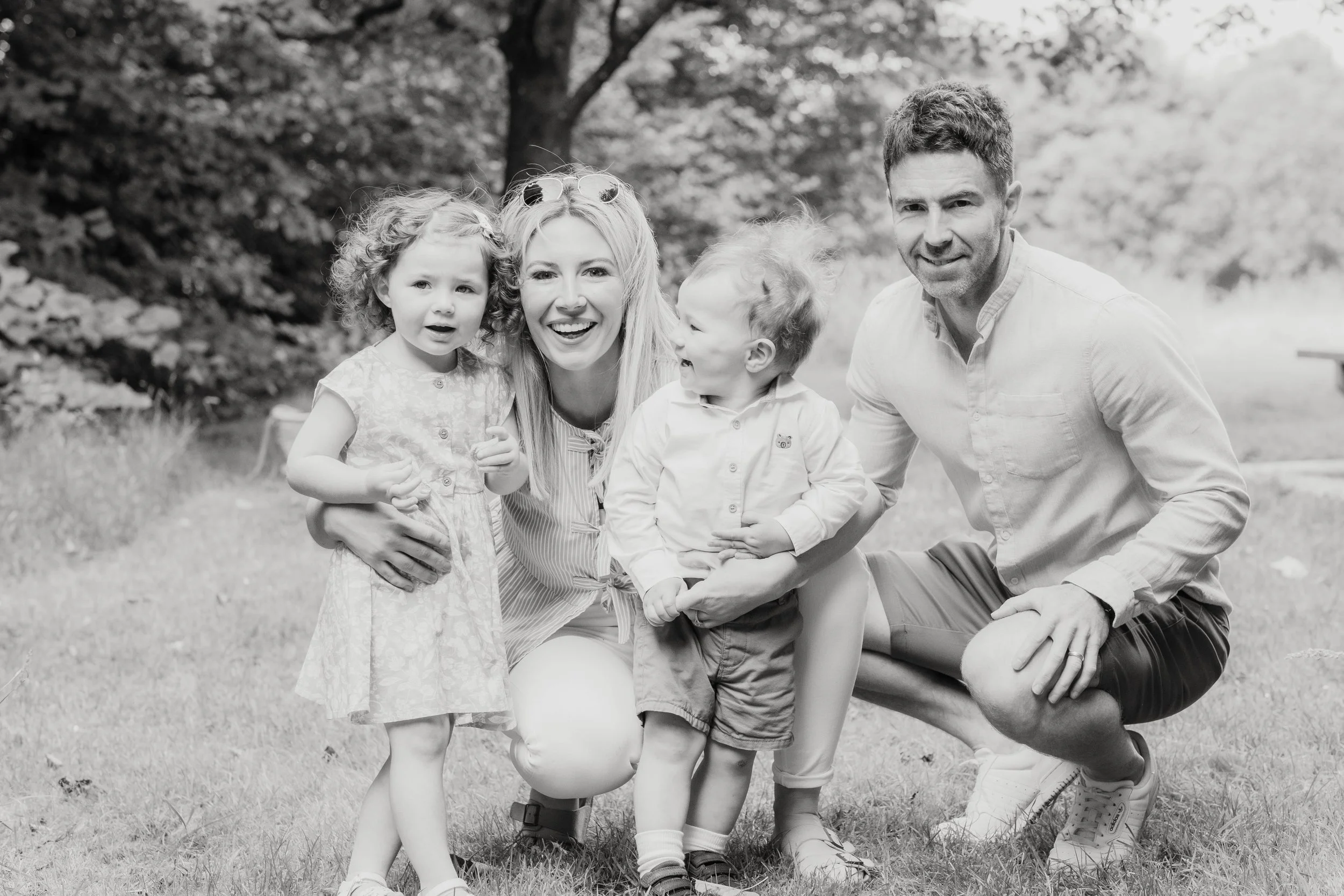 A happy family of four outdoors on a grassy area with trees in the background, including two young children, a woman, and a man, all smiling and posing together.