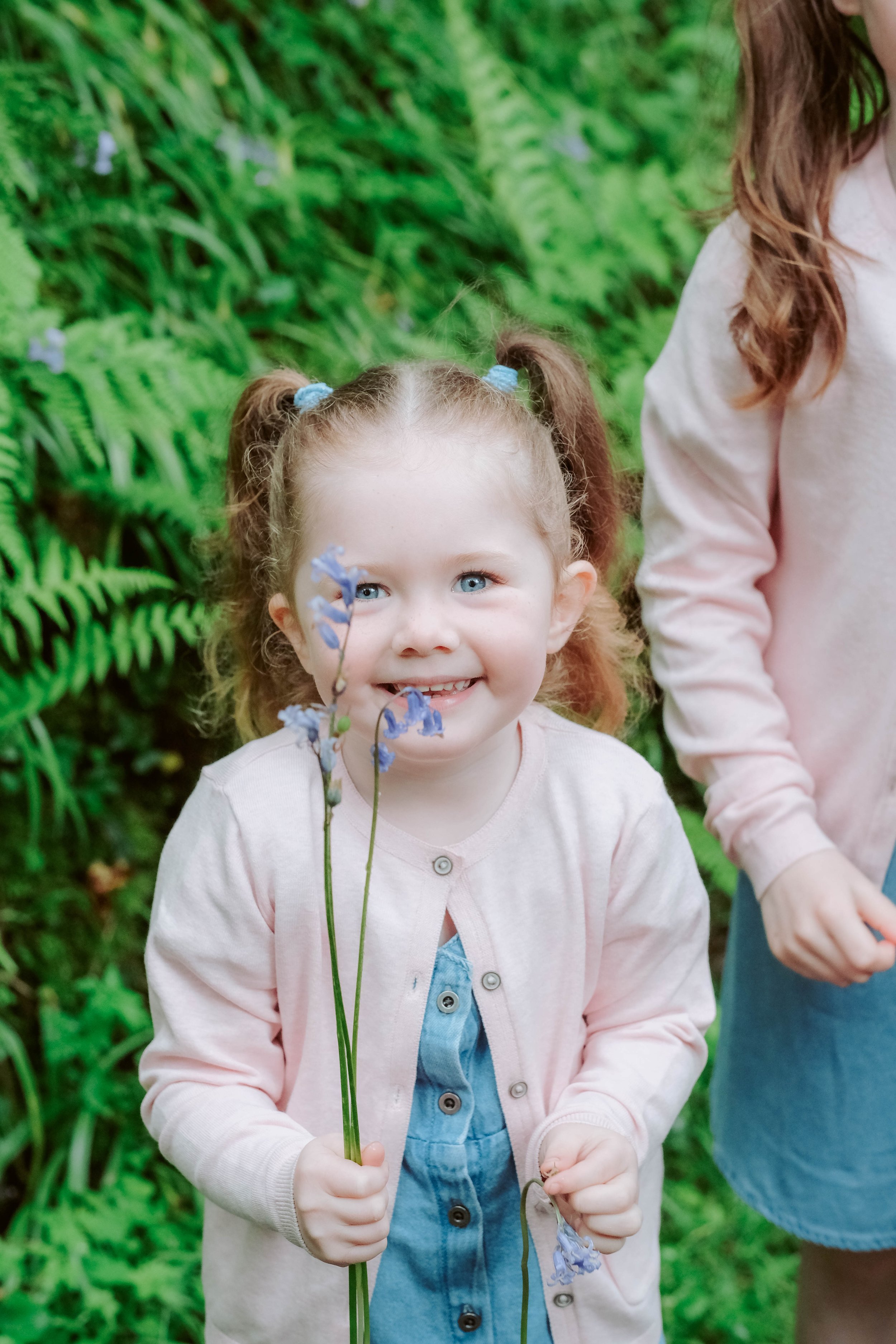A young girl with curly hair in pigtails holding blue flowers and smiling, with greenery in the background.