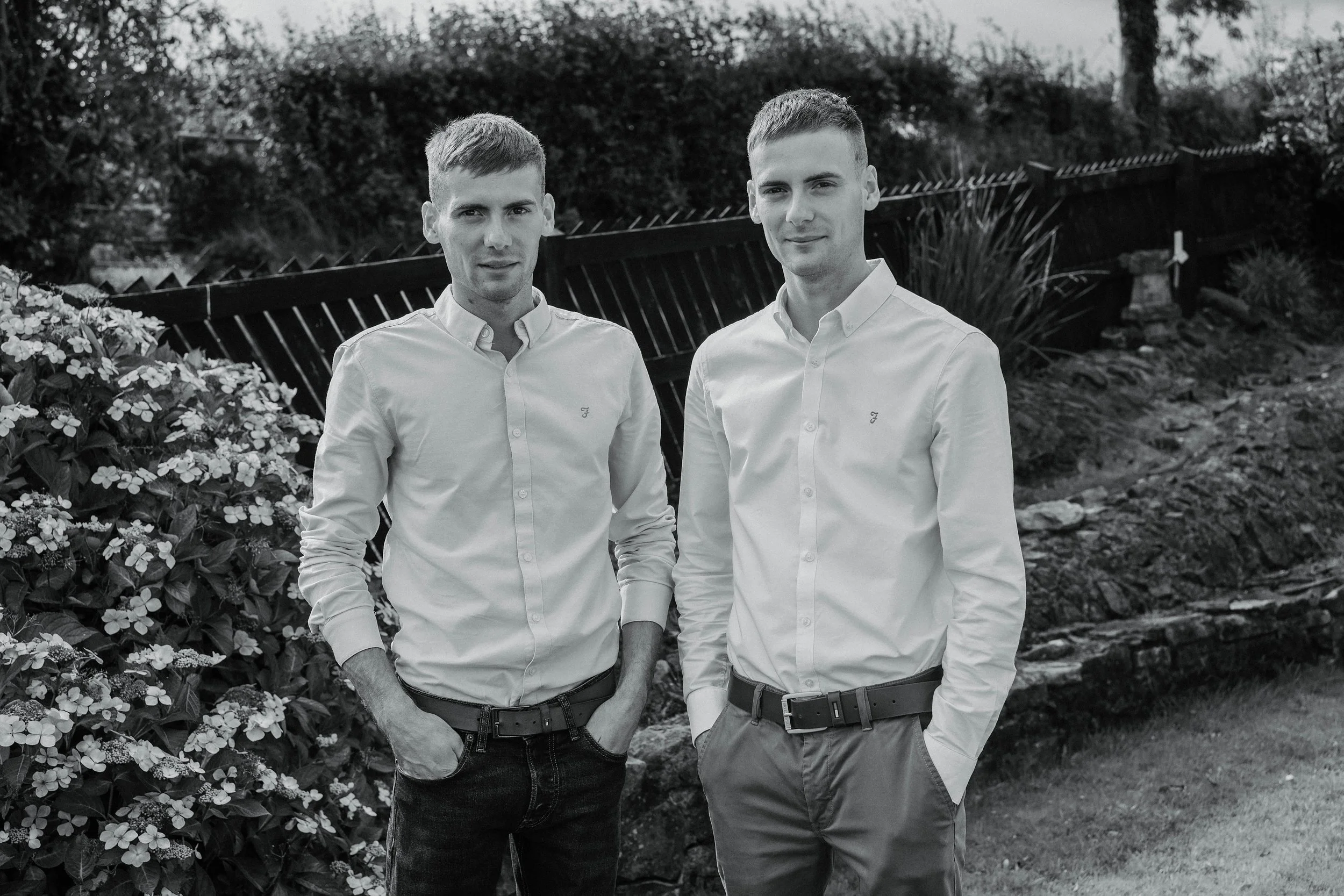 Two young men in white button-up shirts and dark pants standing outdoors in front of a wooden fence and garden.