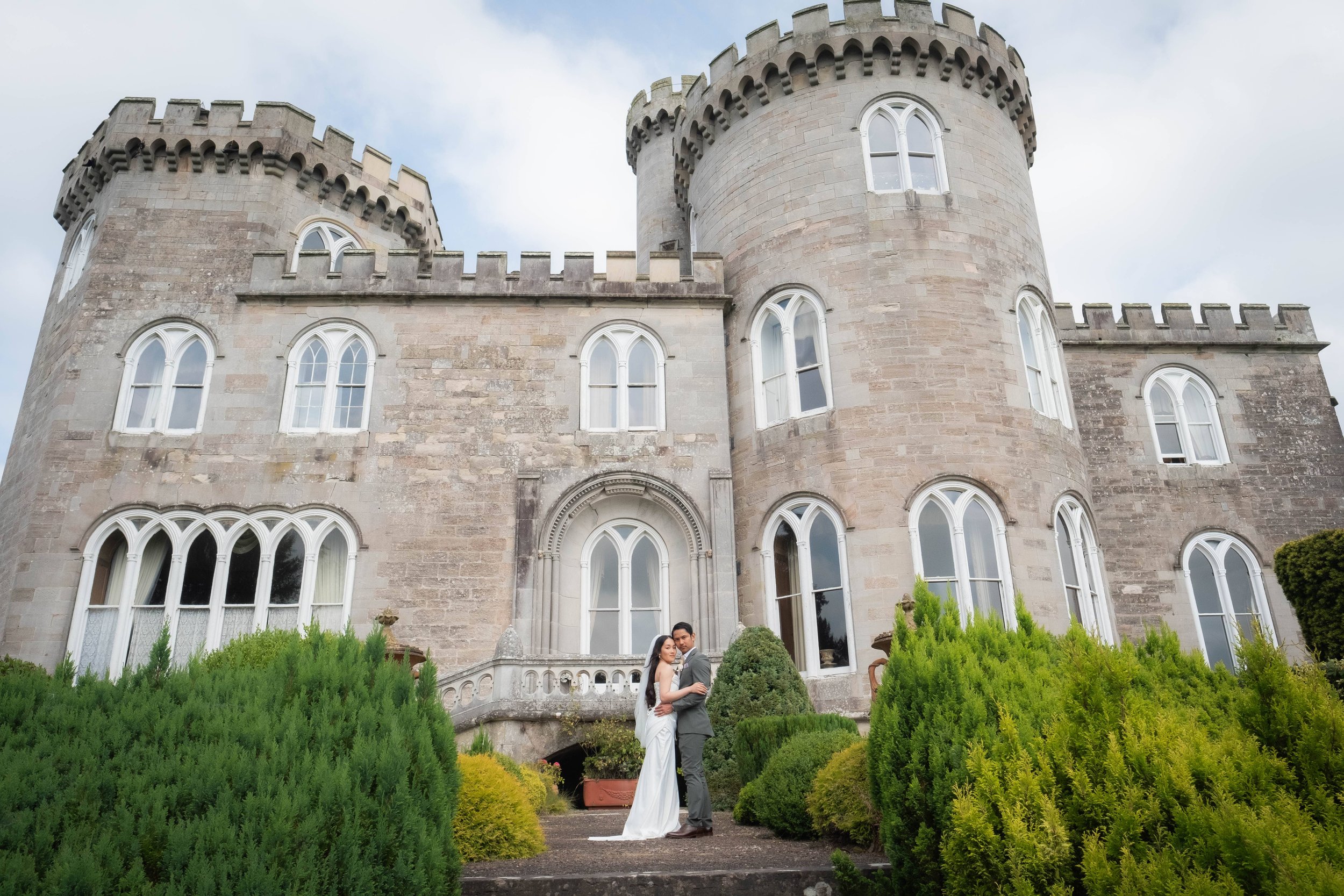 A bride and groom standing close together in front of a large stone castle with multiple tall, arched windows and conical towers. Green bushes and trees surround them.