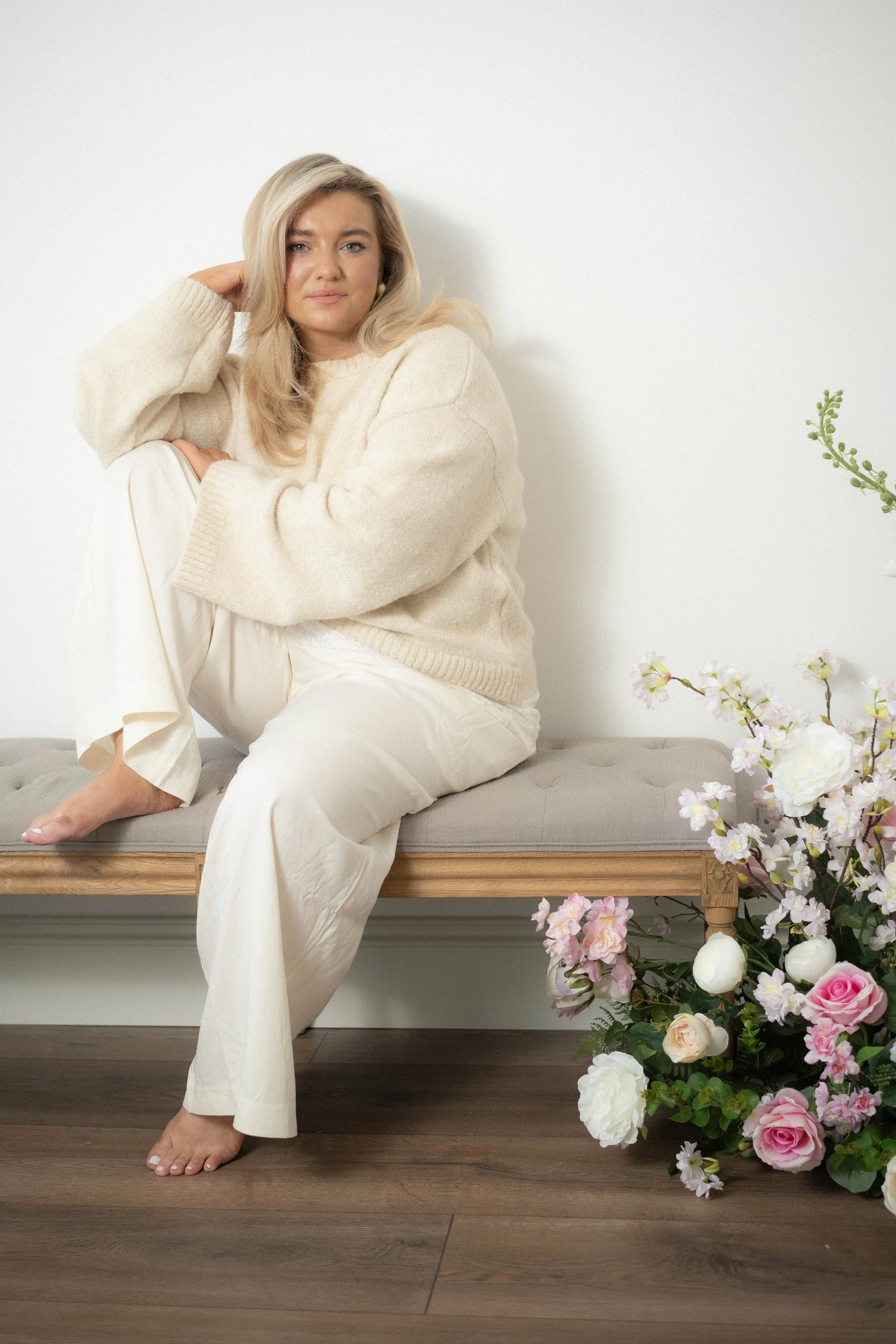A woman with blonde hair sitting on a bench against a white wall, wearing a beige sweater and white pants, with flowers nearby.