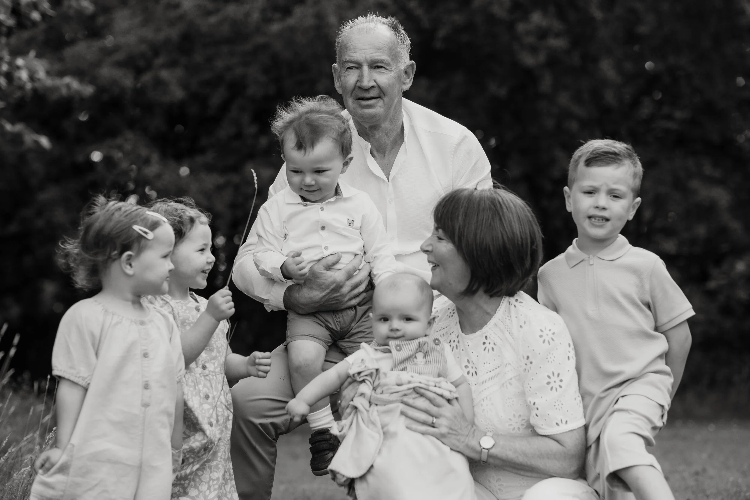 A black-and-white photo of three generations of a family outdoors, including two elderly adults, three young children, and one baby, all engaging and smiling.