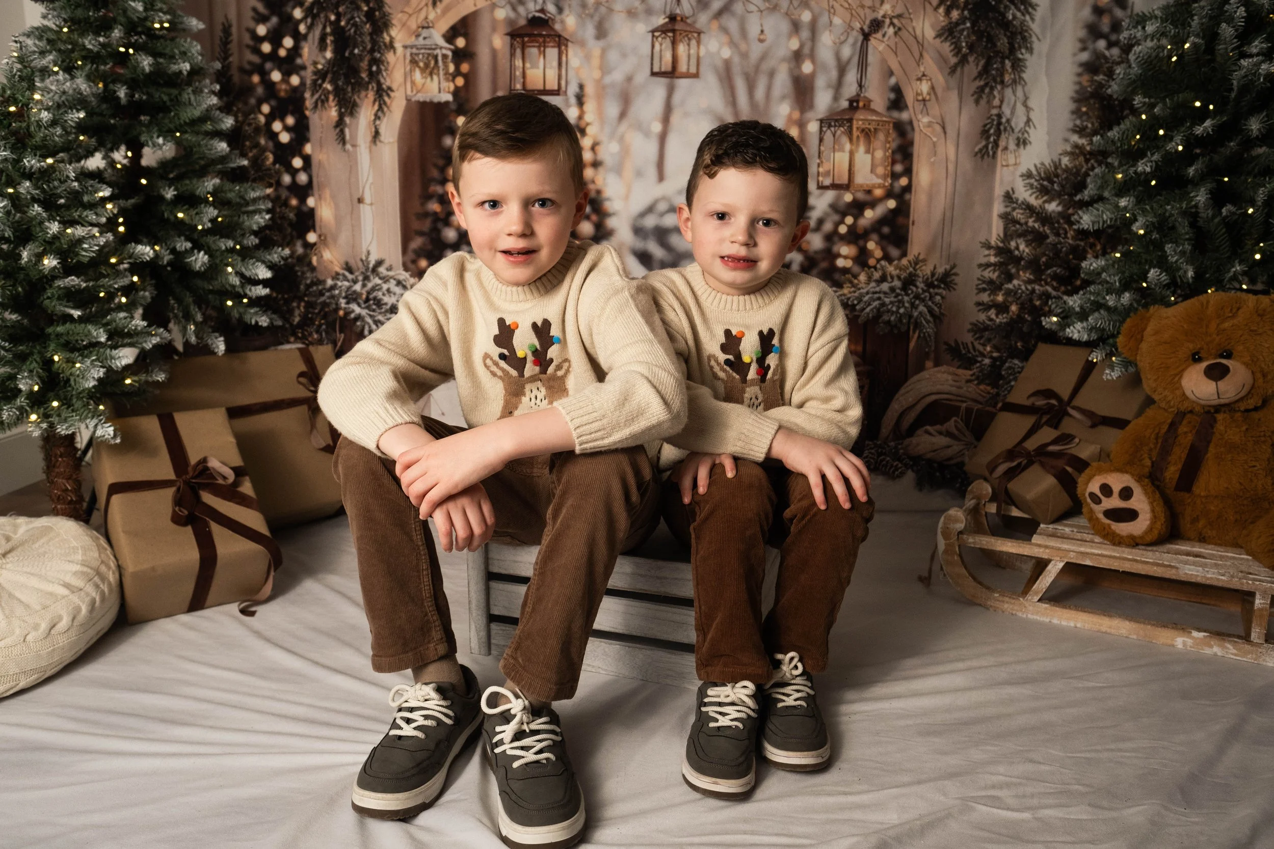Two young boys sitting on a small bench in front of a Christmas-themed backdrop with decorated trees, wrapped presents, and a teddy bear on a wooden sleigh, all in a cozy holiday setting.