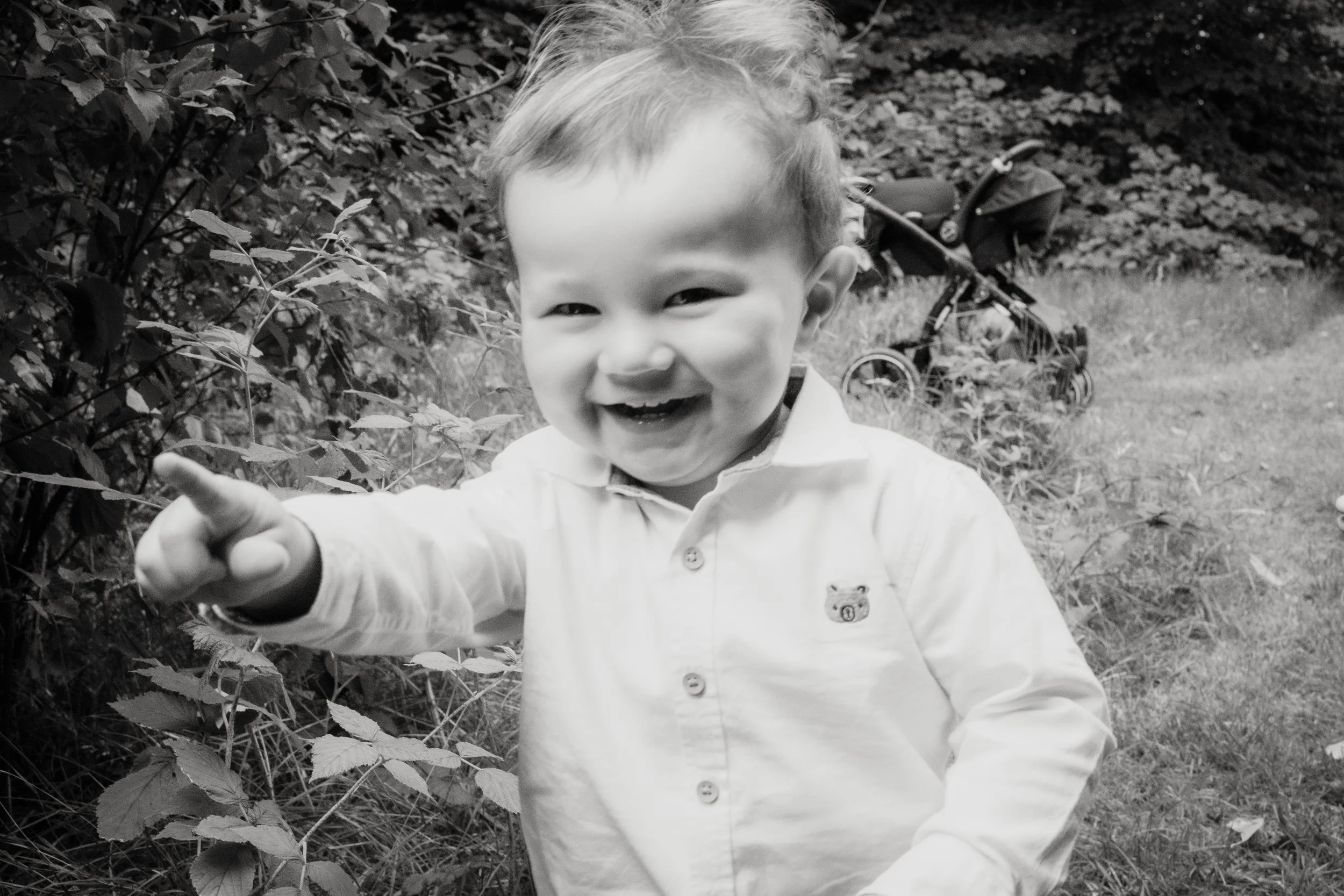 A young boy smiling and pointing at the camera outdoors, with a toy stroller in the background.