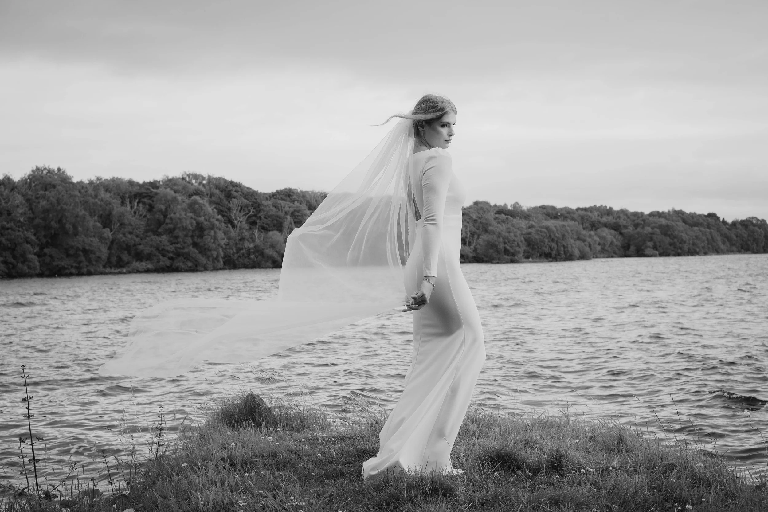 A woman in a wedding dress standing on a grassy shore near a body of water with trees in the background, captured in black and white.