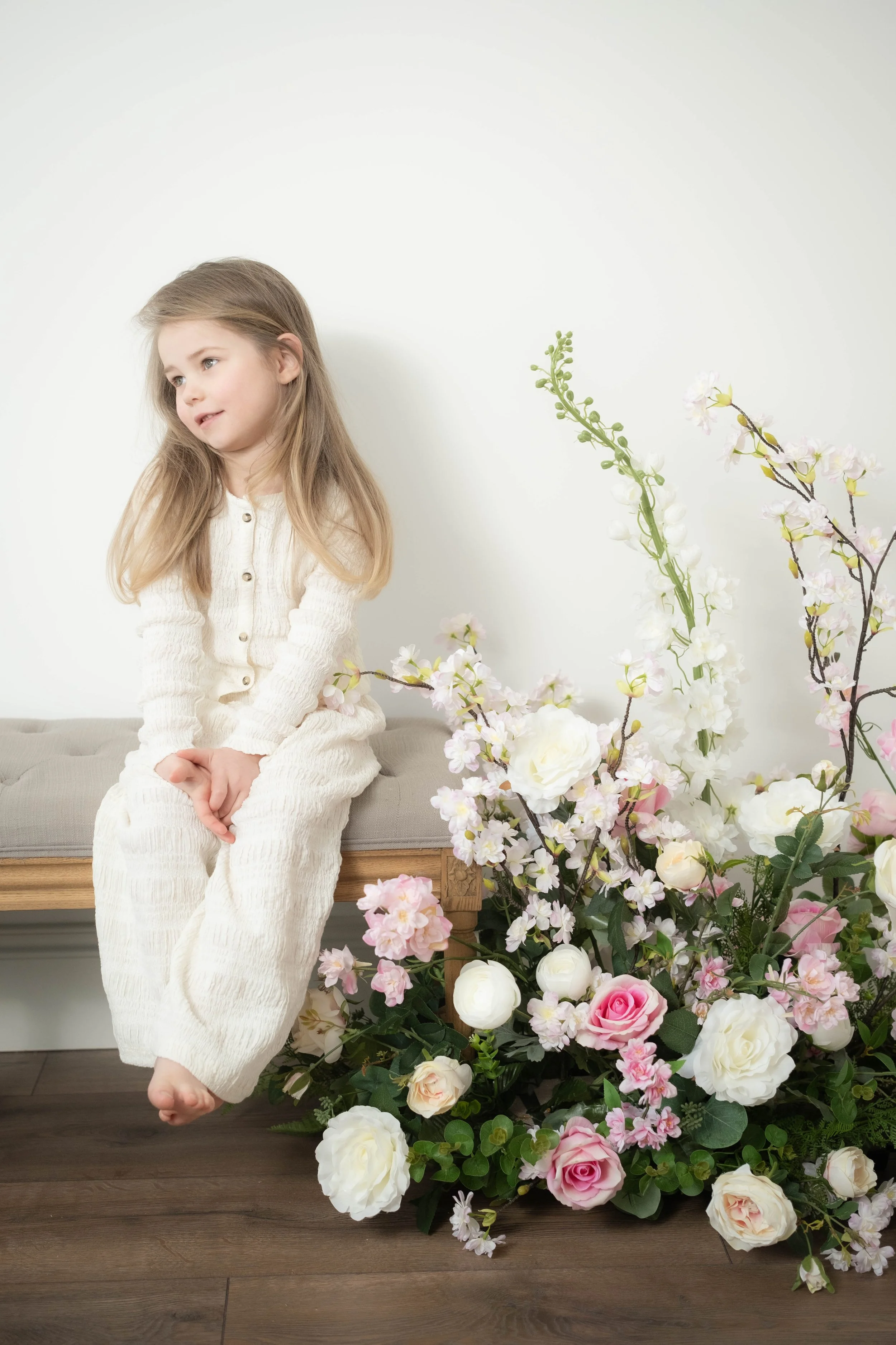A young girl with long blonde hair is sitting on a beige bench, wearing a cream-colored sweater and pants, next to a large arrangement of pink and white flowers against a white wall.