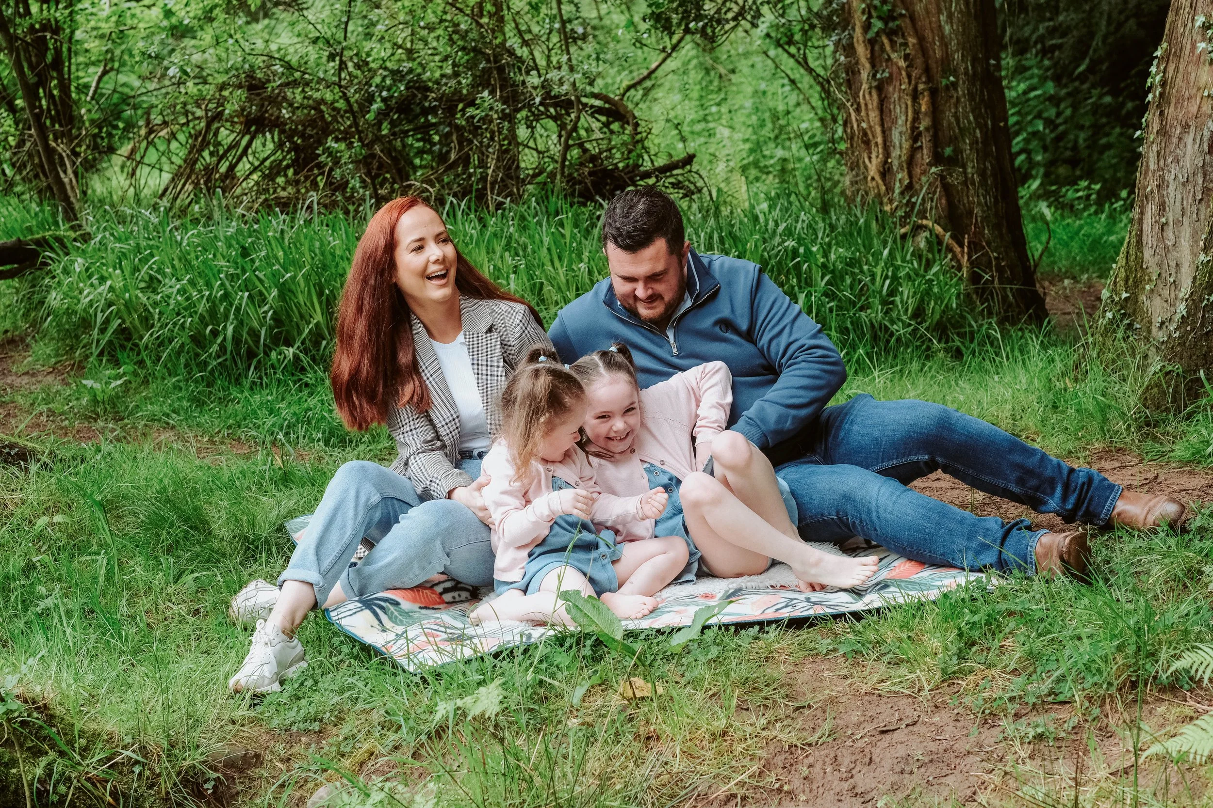 A family of four sitting on a blanket in a forest, laughing and enjoying their time together.