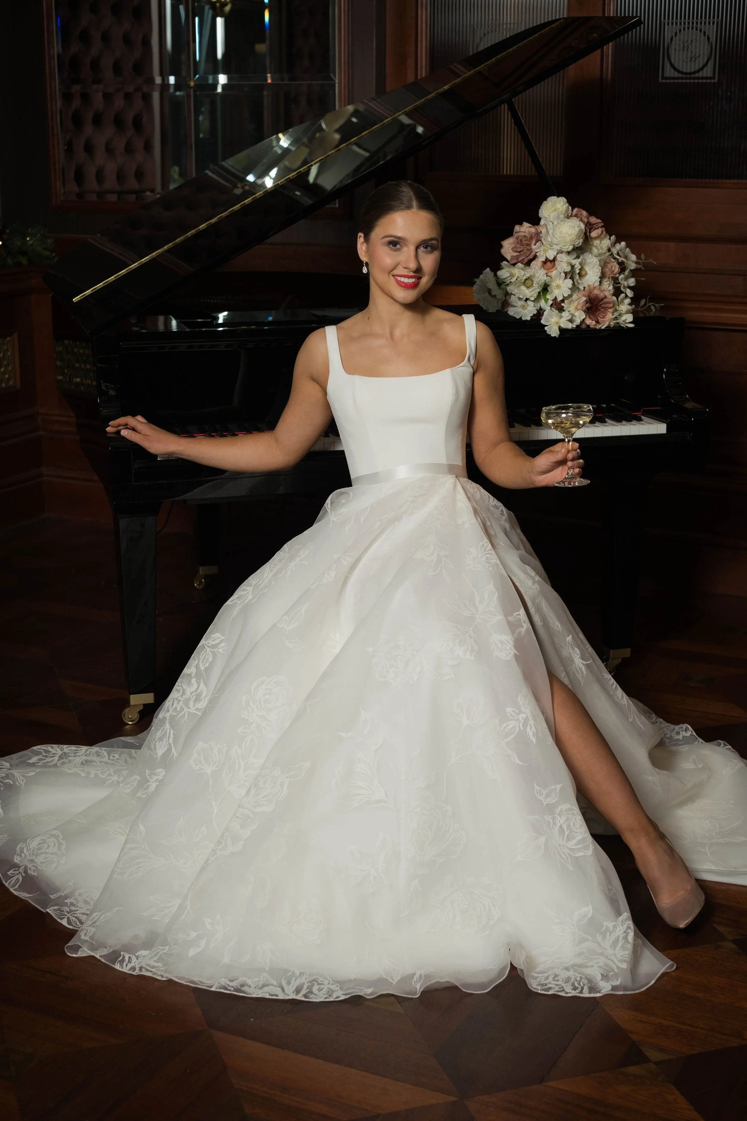 Woman in a white wedding dress sits by a grand piano, smiling at the camera as a wedding photographer captures the moment.