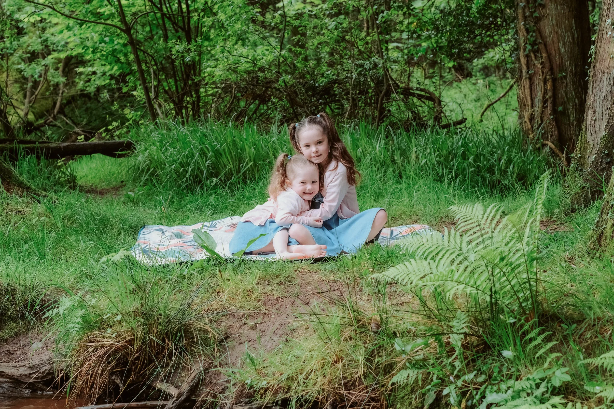 Two young girls, one older with long brown hair and one younger with curly red hair, sitting on a blanket in a lush green forest, hugging and smiling.
