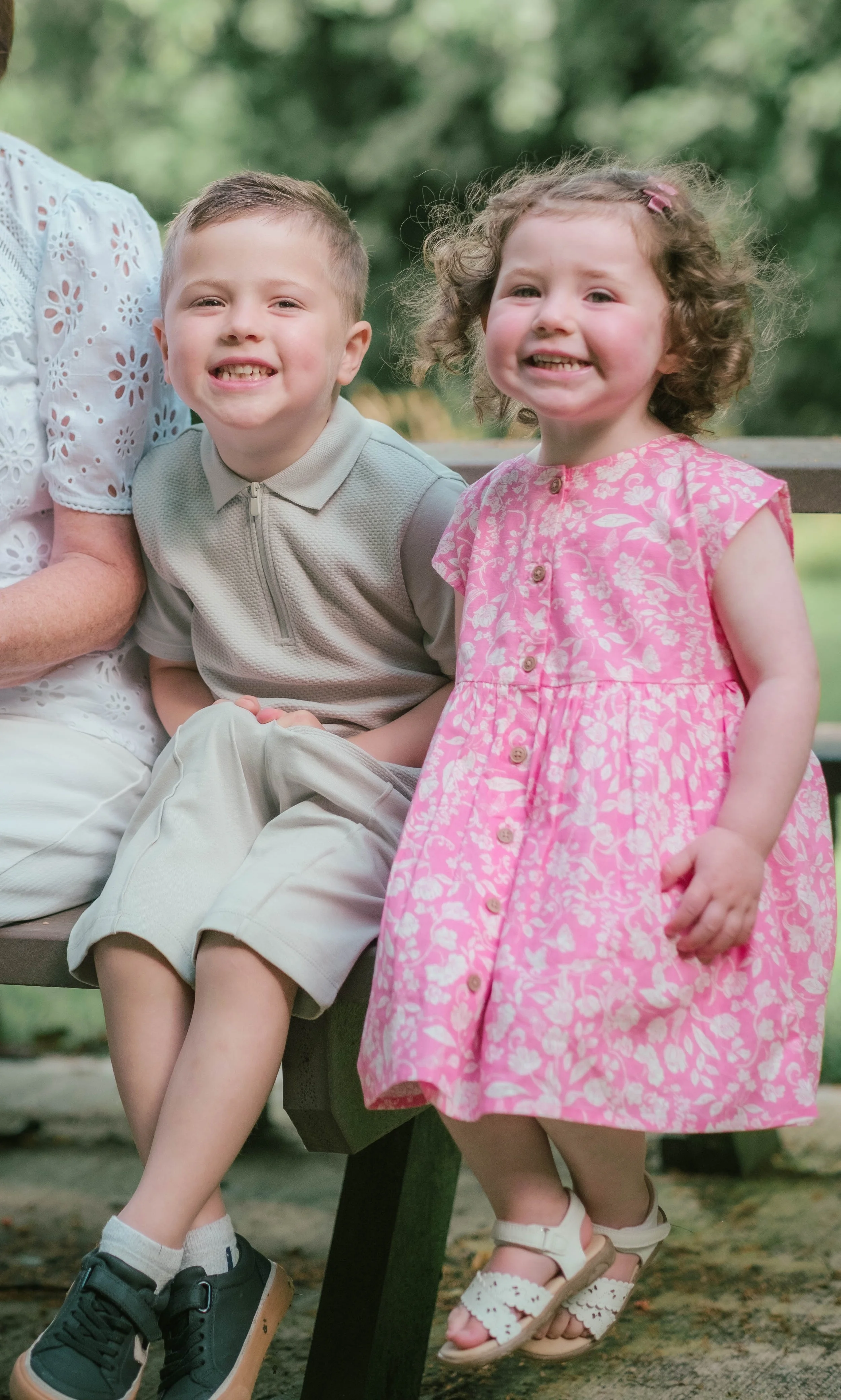 Two smiling children, a boy and a girl, sitting on a park bench outdoors, with a blurred green background.