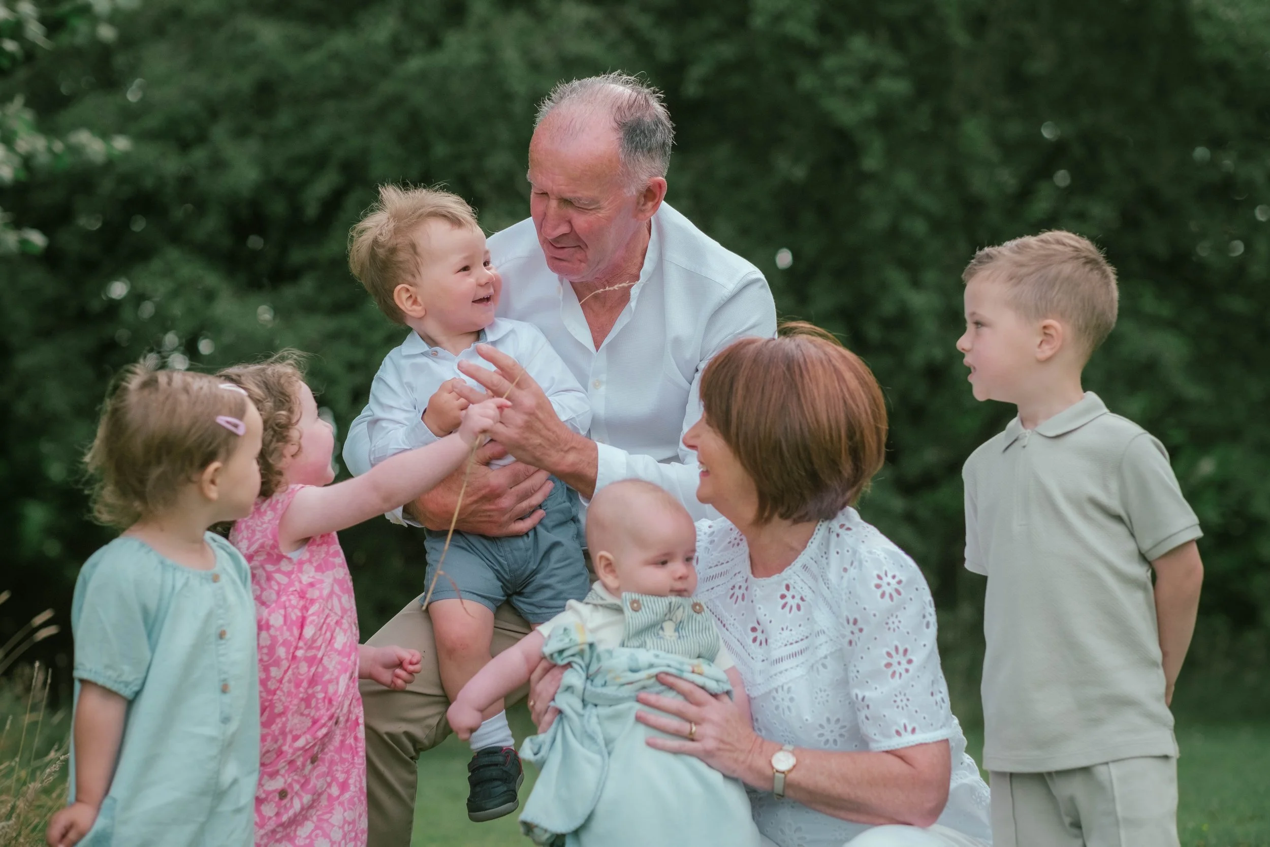 Family gathering outdoors with three children, one women, and one elderly man, enjoying a cheerful moment in a green park.