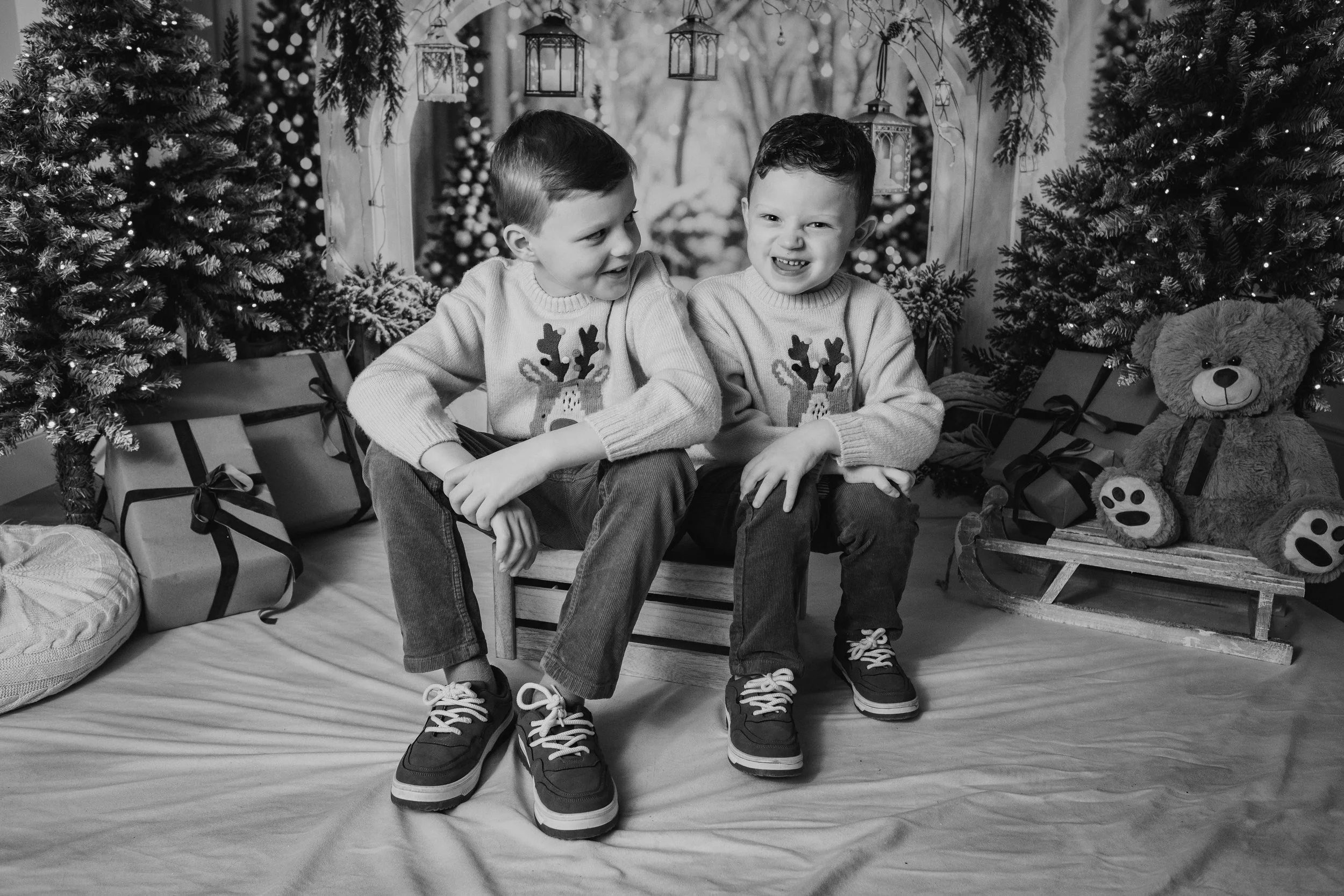 Two young boys sitting on a small bench, wearing Christmas sweaters with reindeer designs, surrounded by decorated Christmas trees, presents, and a teddy bear on a sled in a festive holiday setting.