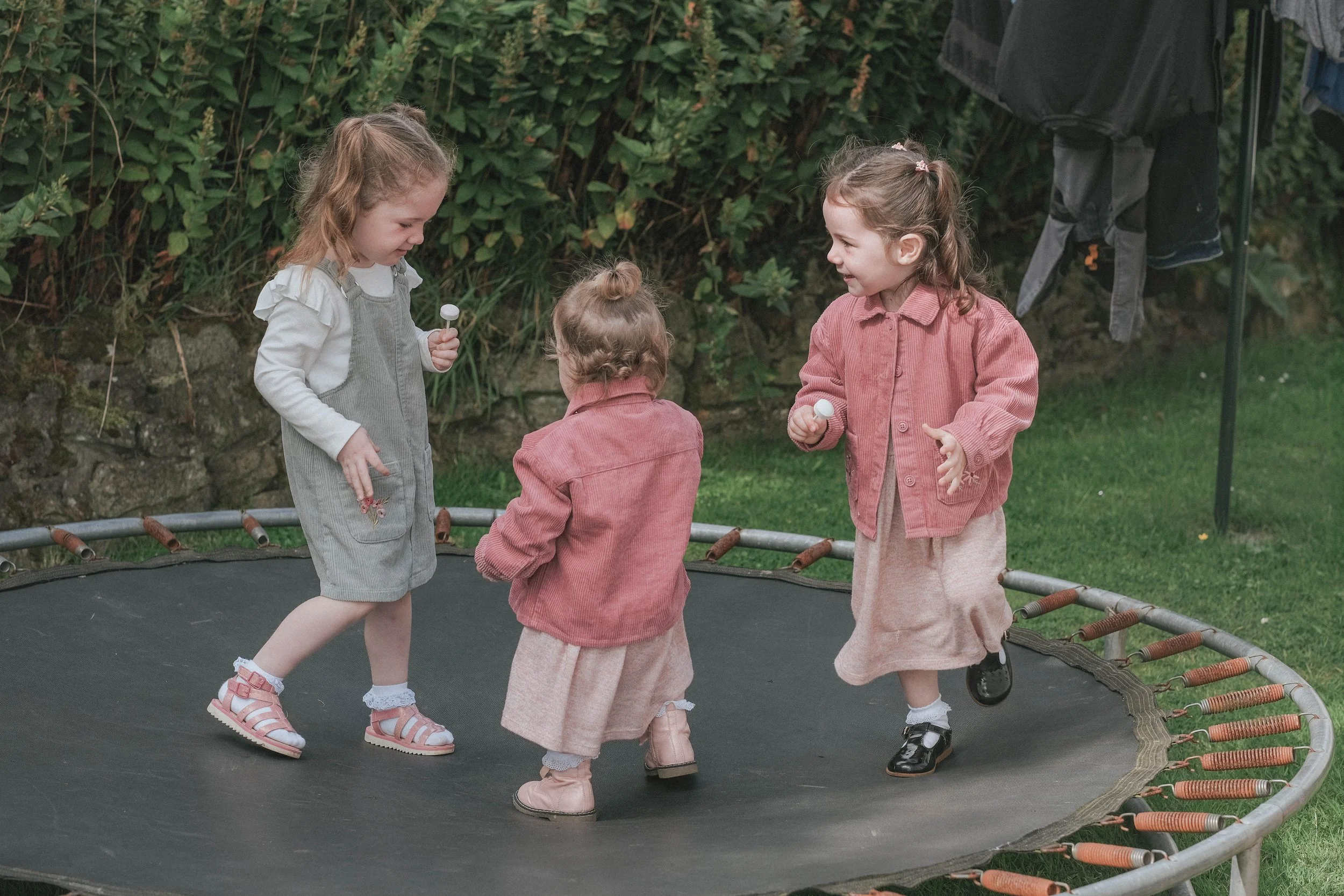Three young girls with light brown hair playing on a small outdoor trampoline, holding small objects in their hands, with a green garden and clothes hanging in the background.