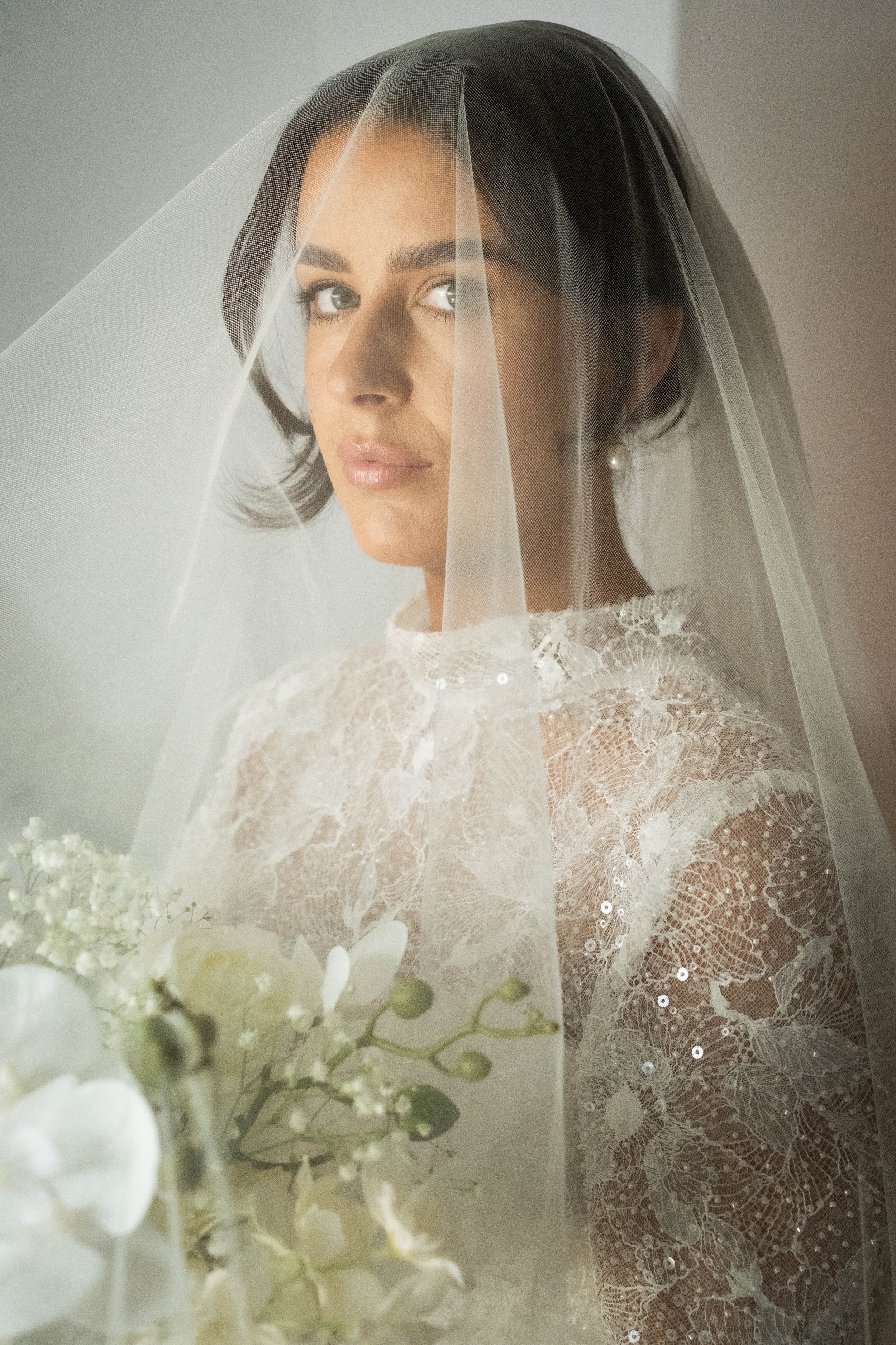 A bride in a lace wedding dress holds a bouquet, gazing forward with her veil down—captured by a skilled wedding photographer.