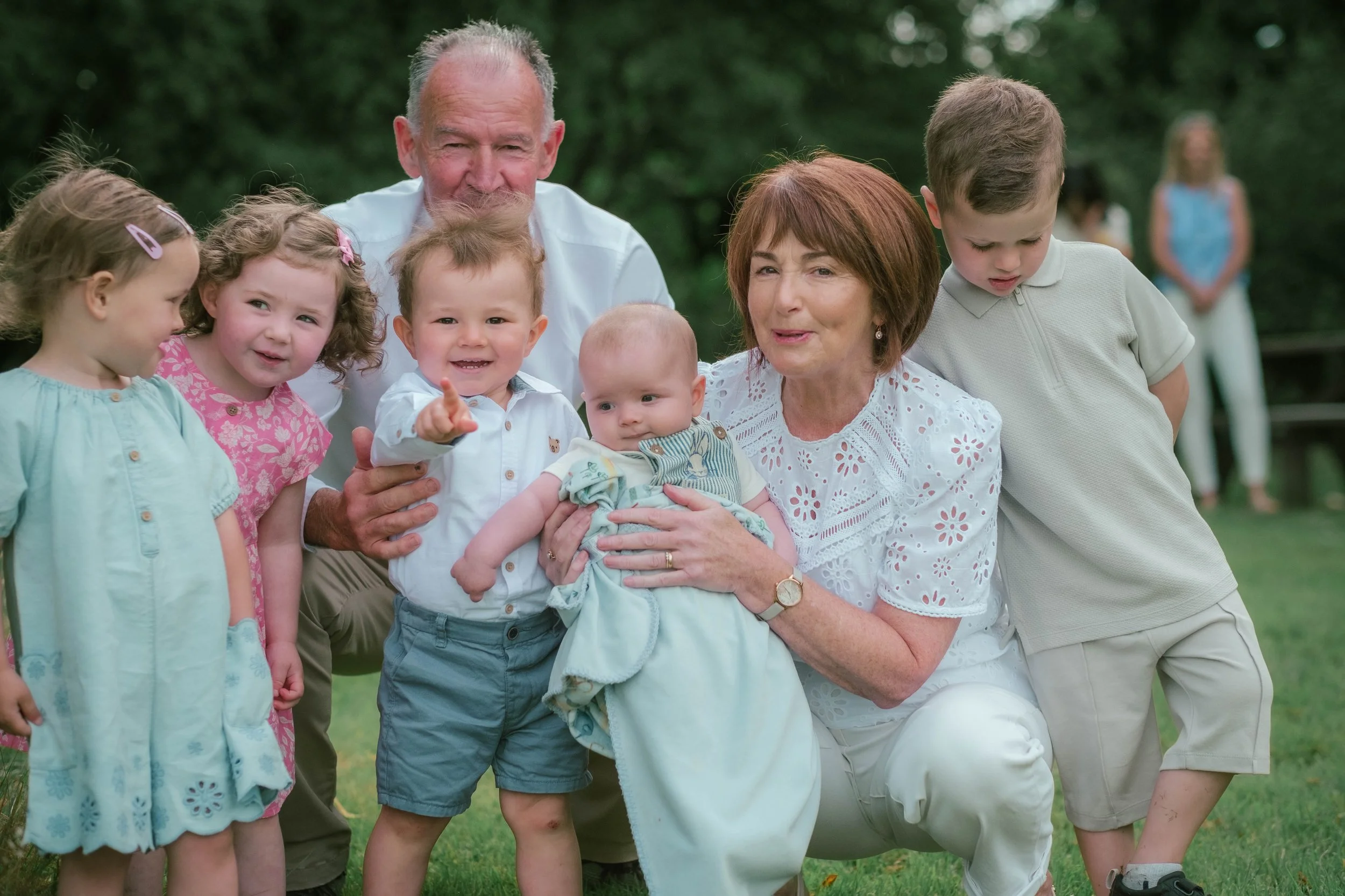 A group of children and elderly adults gathered outdoors for a photo, with some kids smiling and others looking curious, on a grassy area with trees in the background.