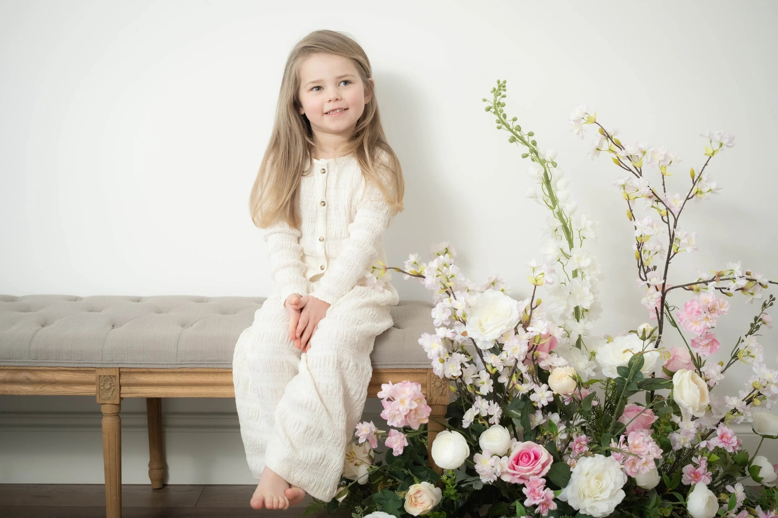 A young girl with long blonde hair sitting on a beige upholstered bench next to a large bouquet of white and pink flowers.