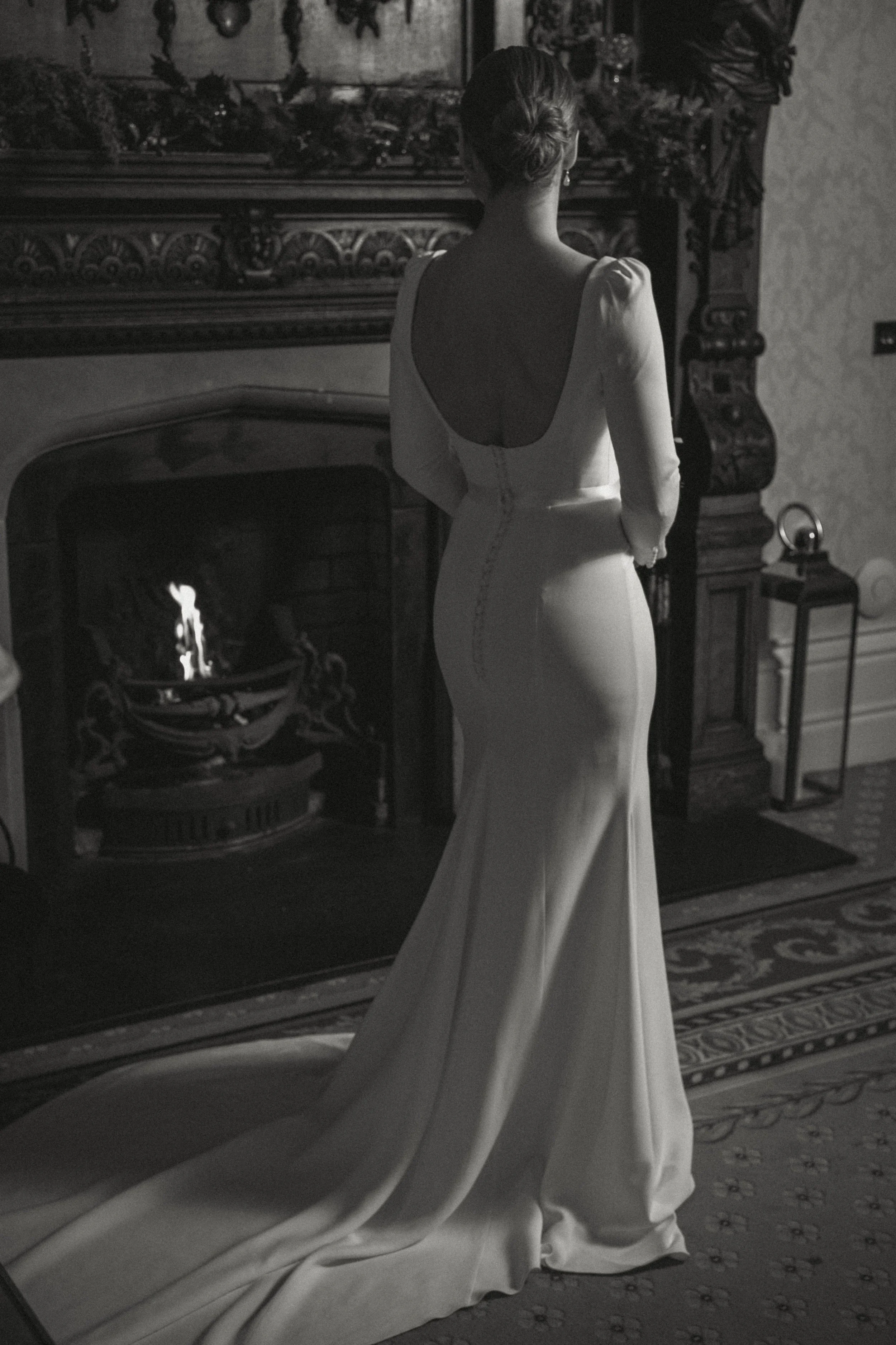 A woman in a long-sleeved wedding dress stands facing a fireplace in an elegant room, her back to the camera.