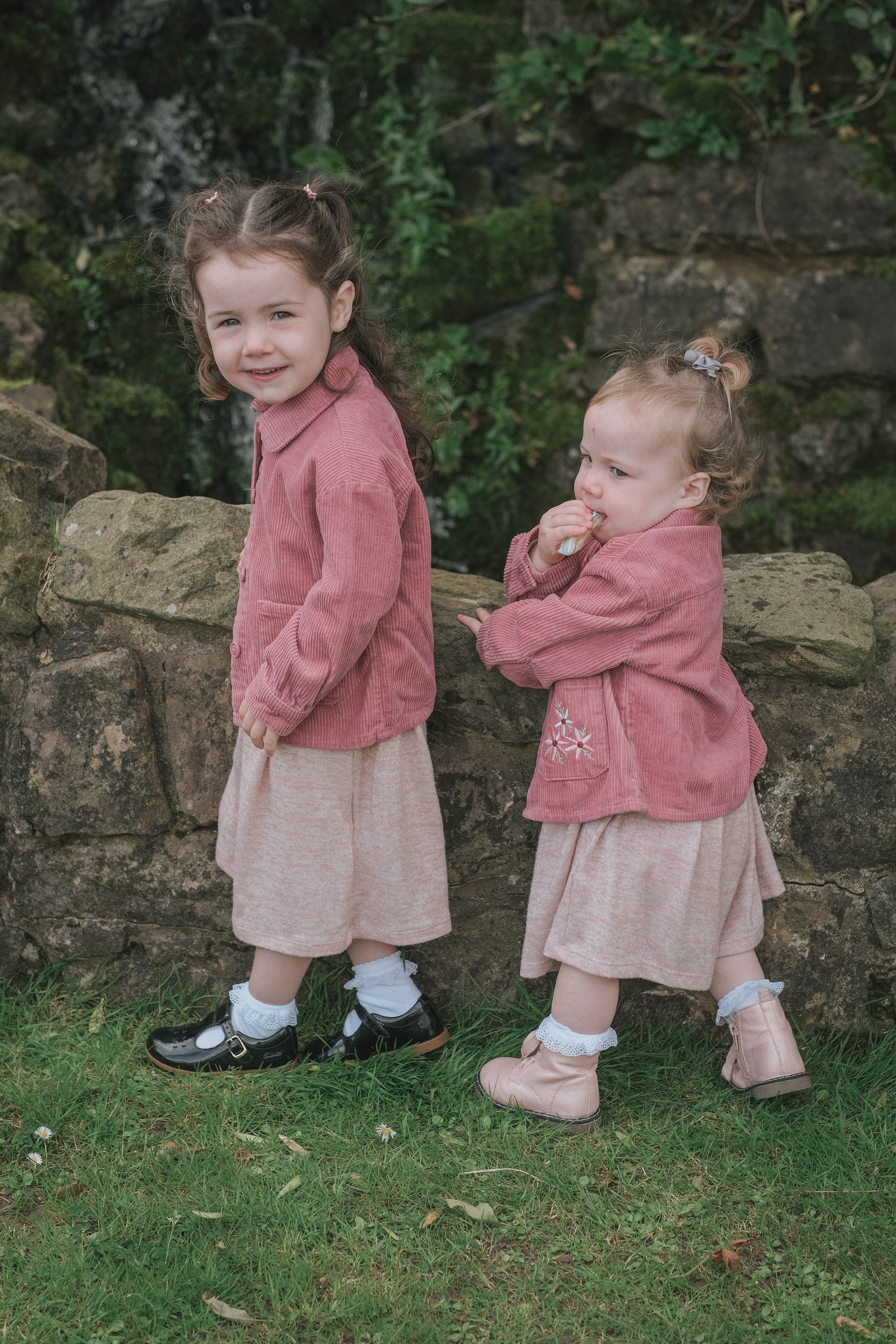 Two young girls with curly hair, wearing pink jackets and beige skirts, standing outdoors on grass in front of a stone wall and greenery.