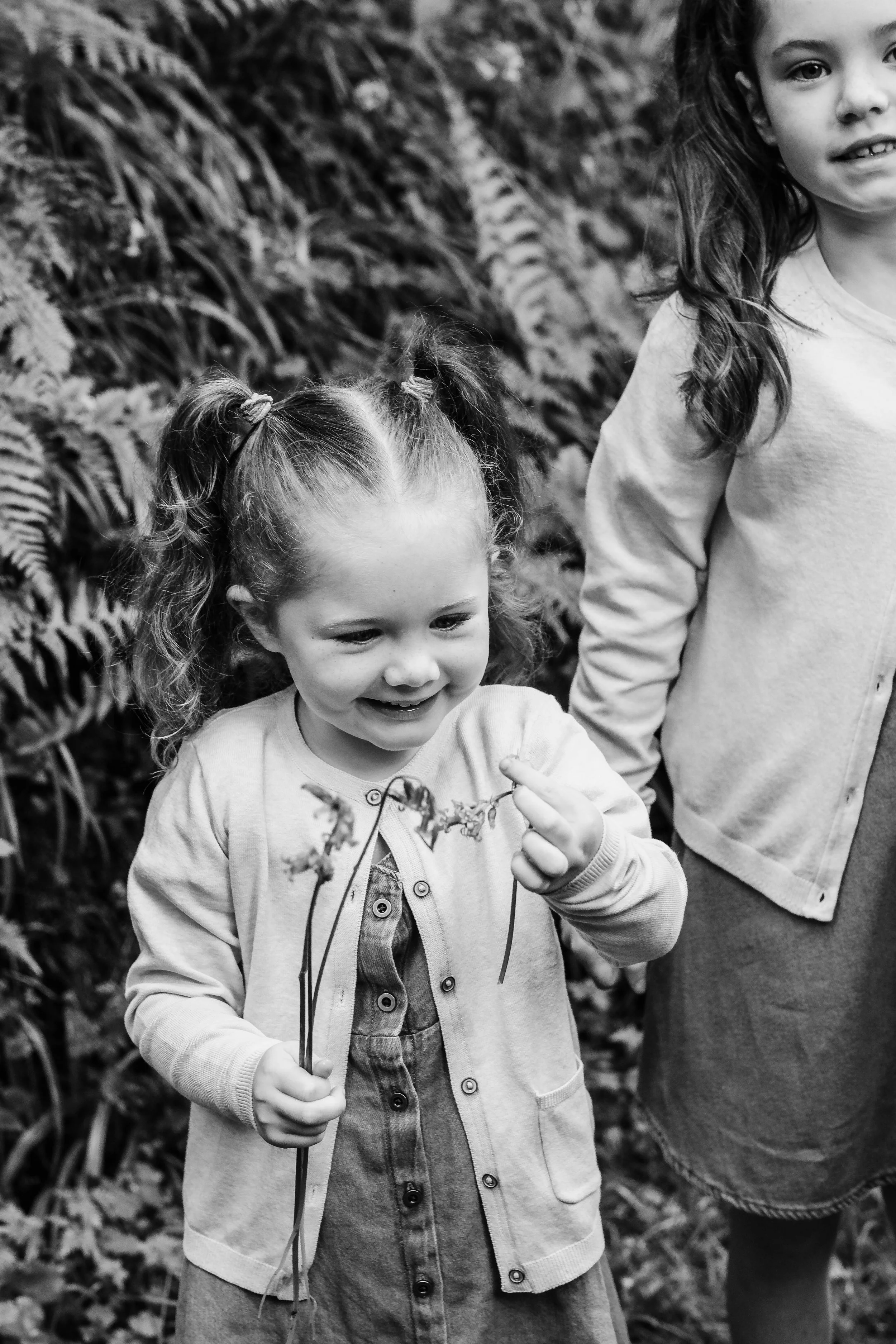 Two young girls outdoors, one smiling and holding a small branch, surrounded by foliage.