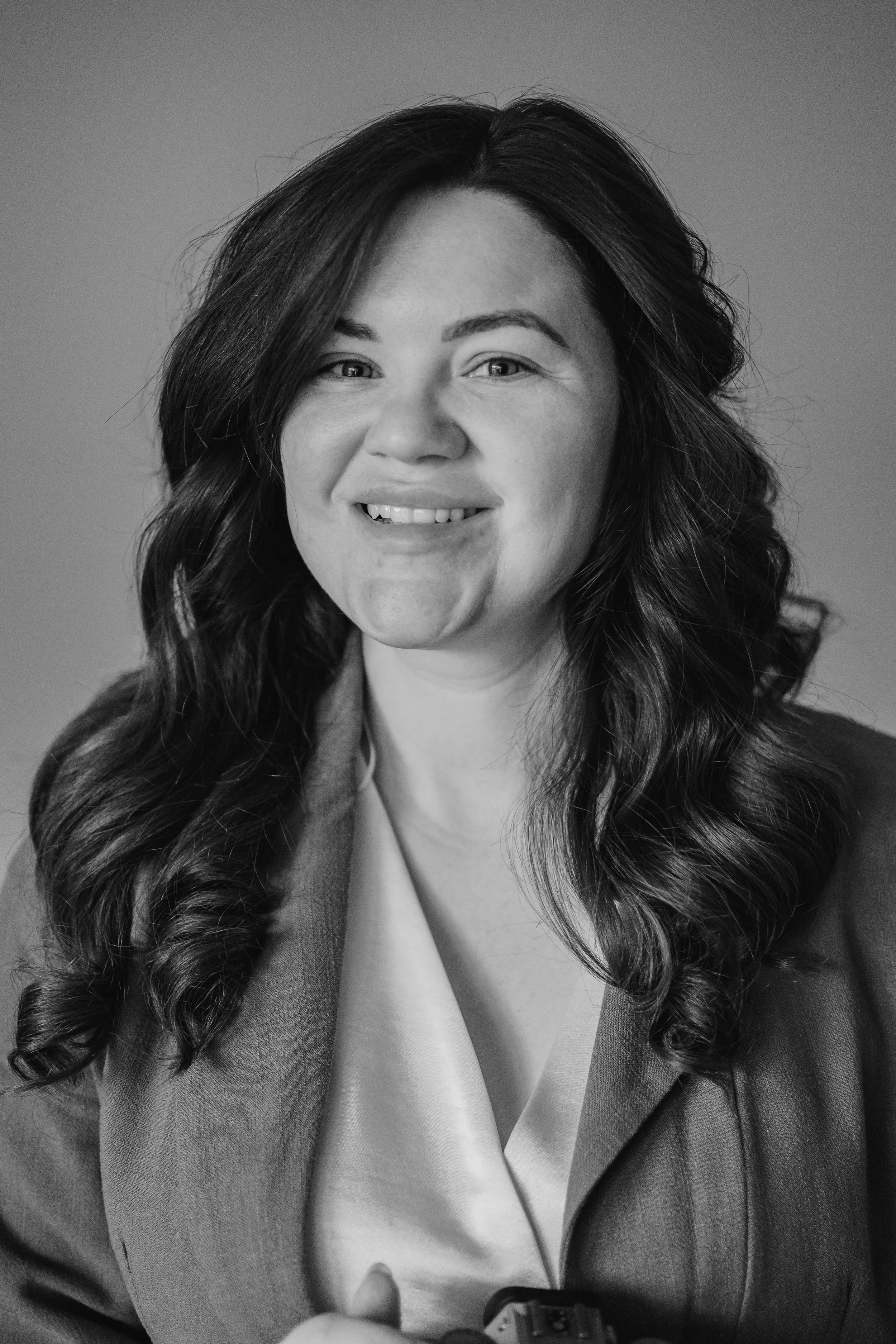 Black and white portrait of a woman with long, wavy hair, smiling and wearing a blazer over a light-colored top.