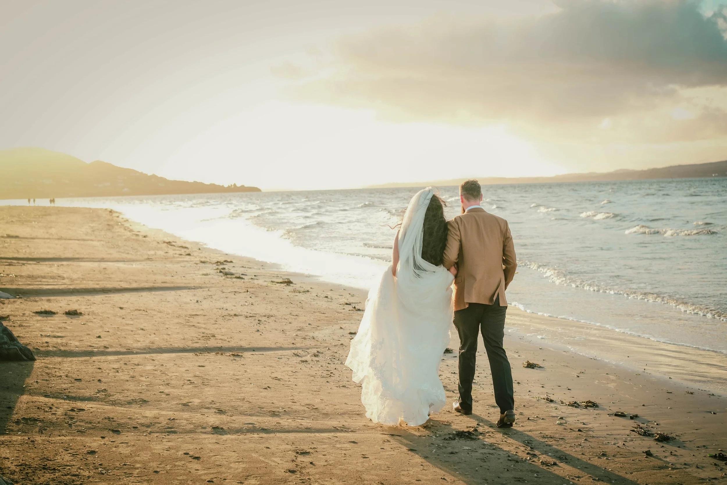 A bride and groom walk along a sandy beach at sunset, holding each other close—perfect for any wedding photographer to capture.