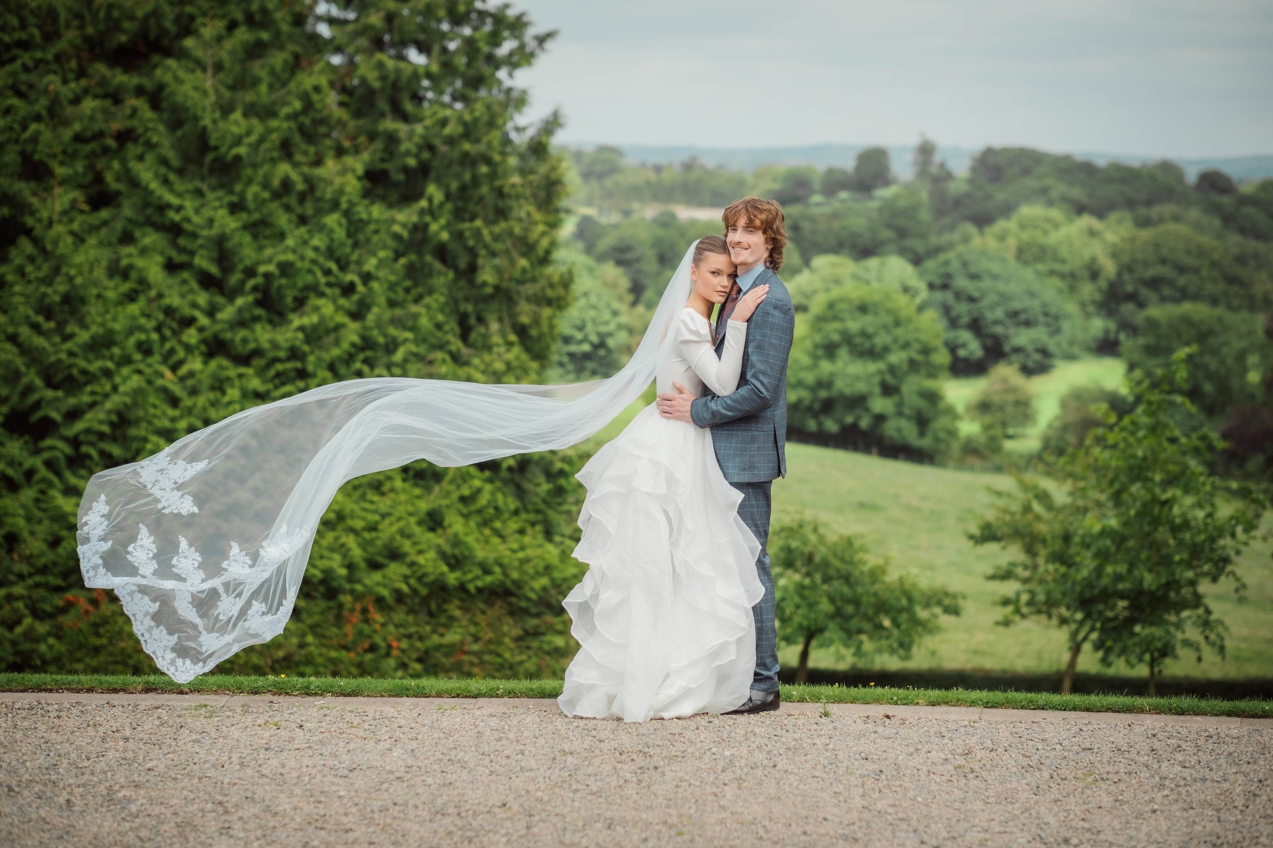 Bride and groom embracing outdoors with green trees and rolling hills in the background.