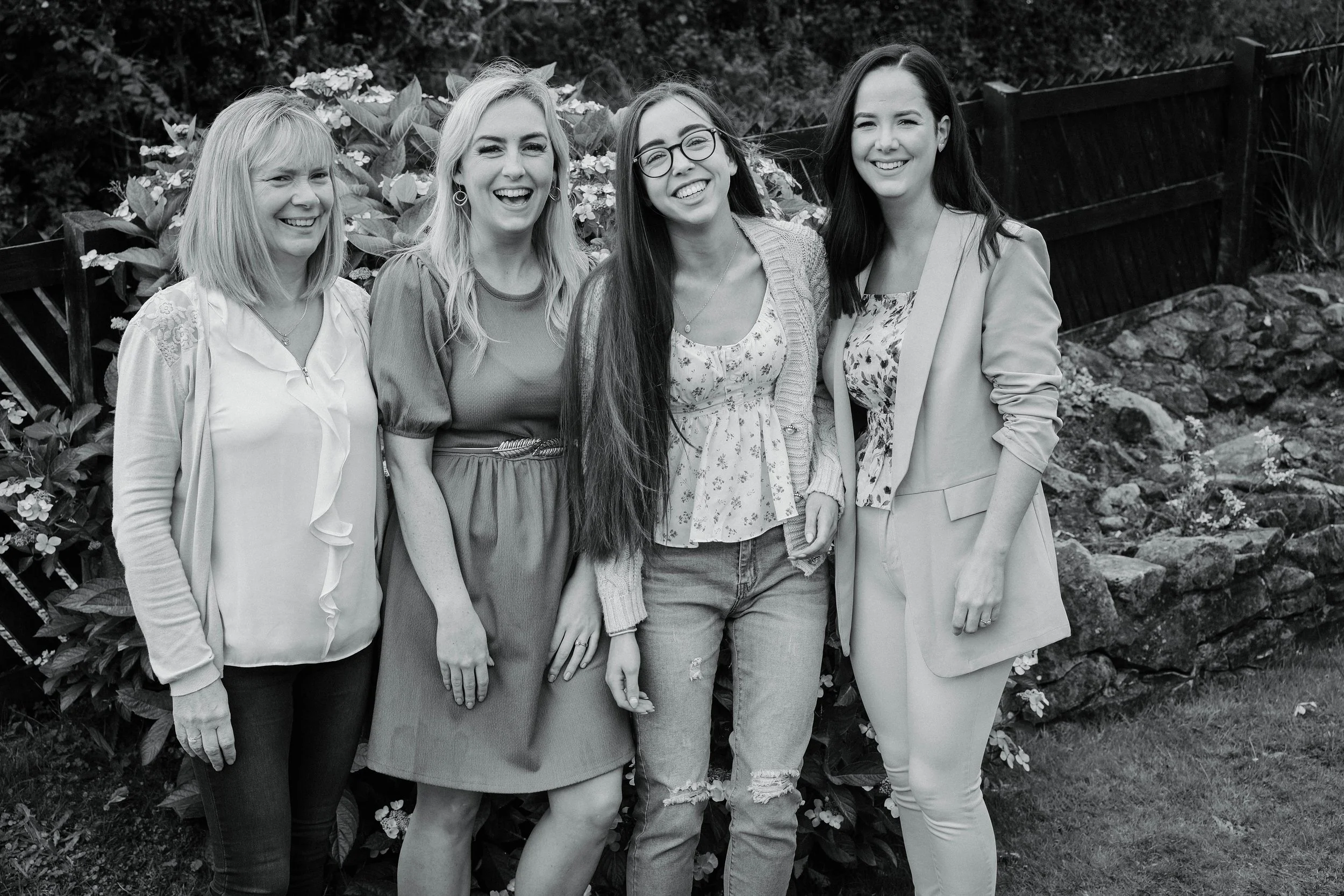 Four women standing outdoors, smiling, with flowers and a garden fence in the background.