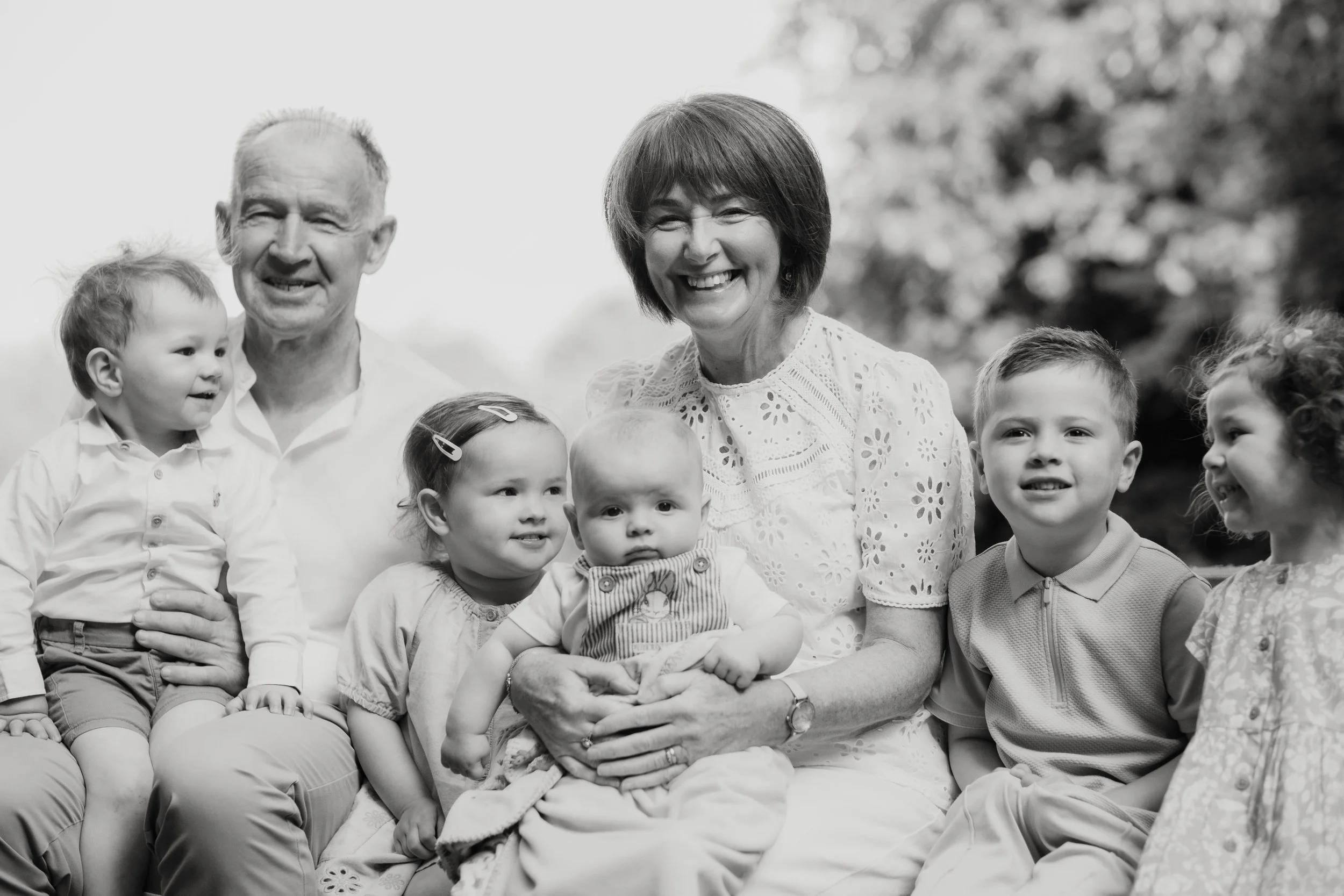 A black-and-white photo of a happy multigenerational family outdoors. The family includes two older adults, a woman and a man, surrounded by five young children. The woman is holding a baby, and everyone is smiling or looking at each other.