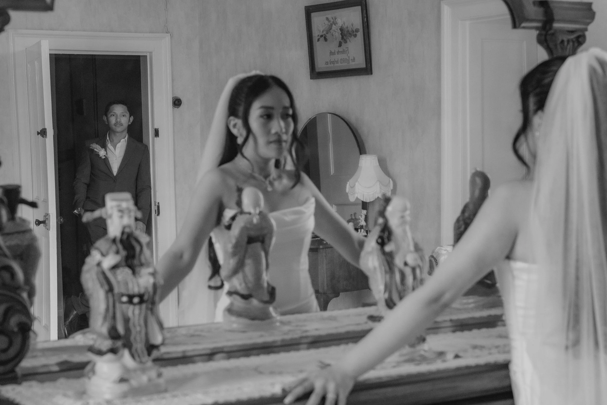 A bride in a white dress looks at herself in a mirror as a man in a suit stands in the doorway behind her.