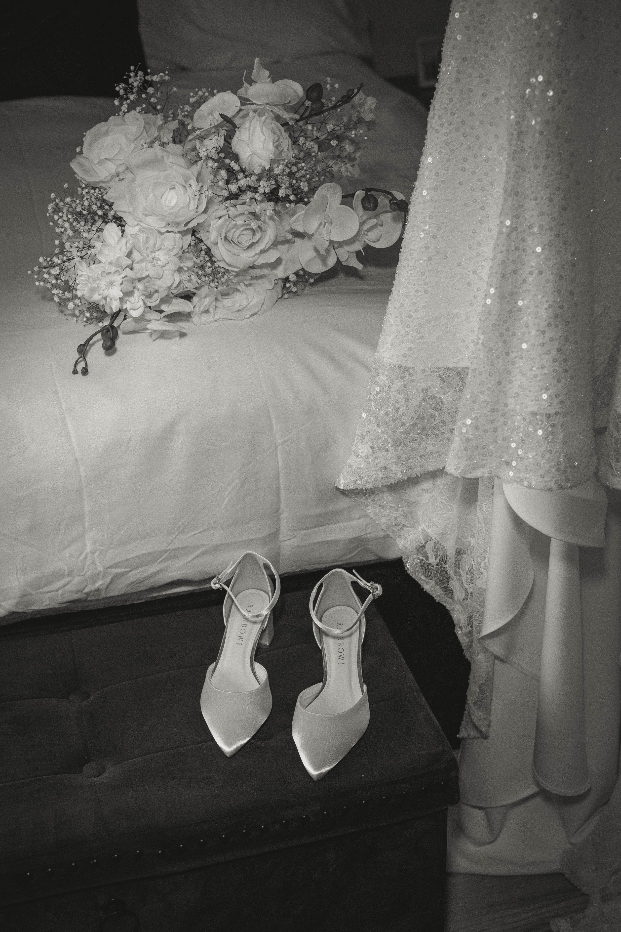 White bridal shoes on a bench, a lace wedding dress, and flowers await the wedding photographer in a softly lit room.