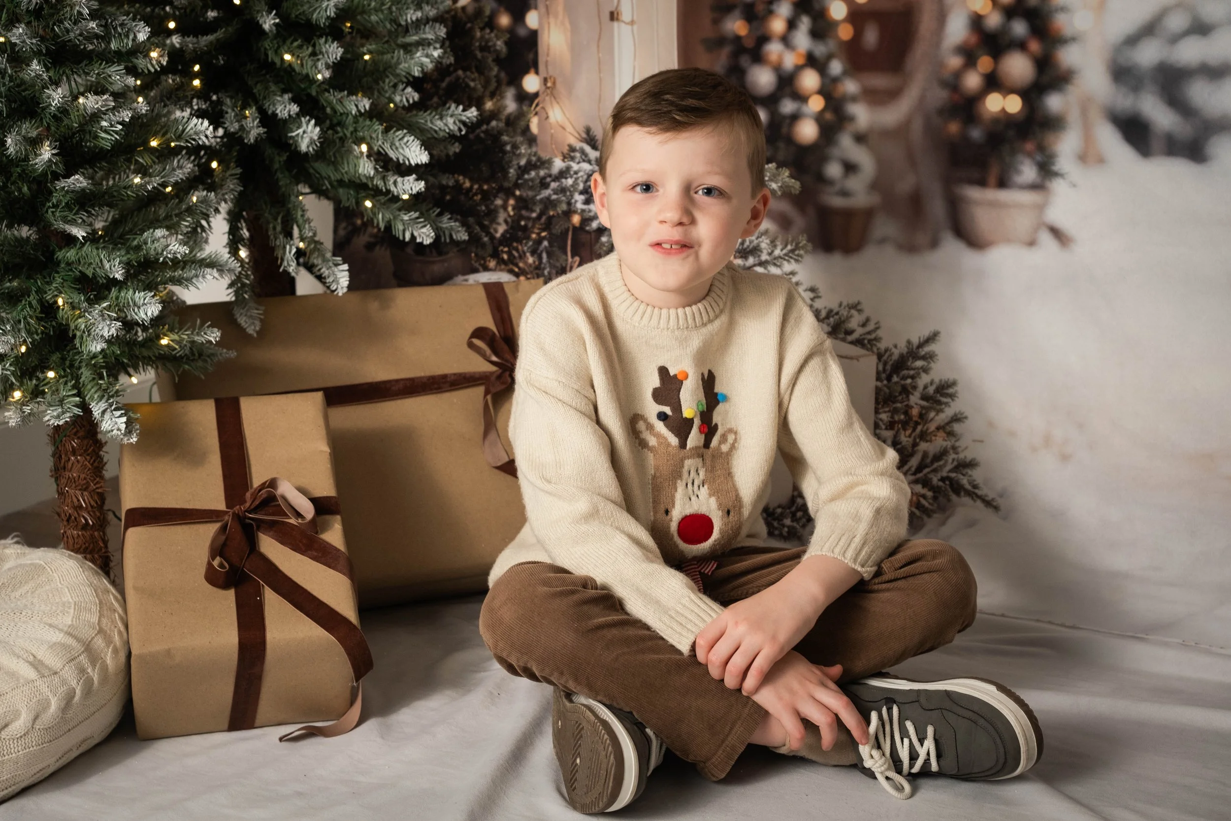 A young boy sitting cross-legged on the floor next to decorated Christmas trees, wrapped presents, and holiday decorations.